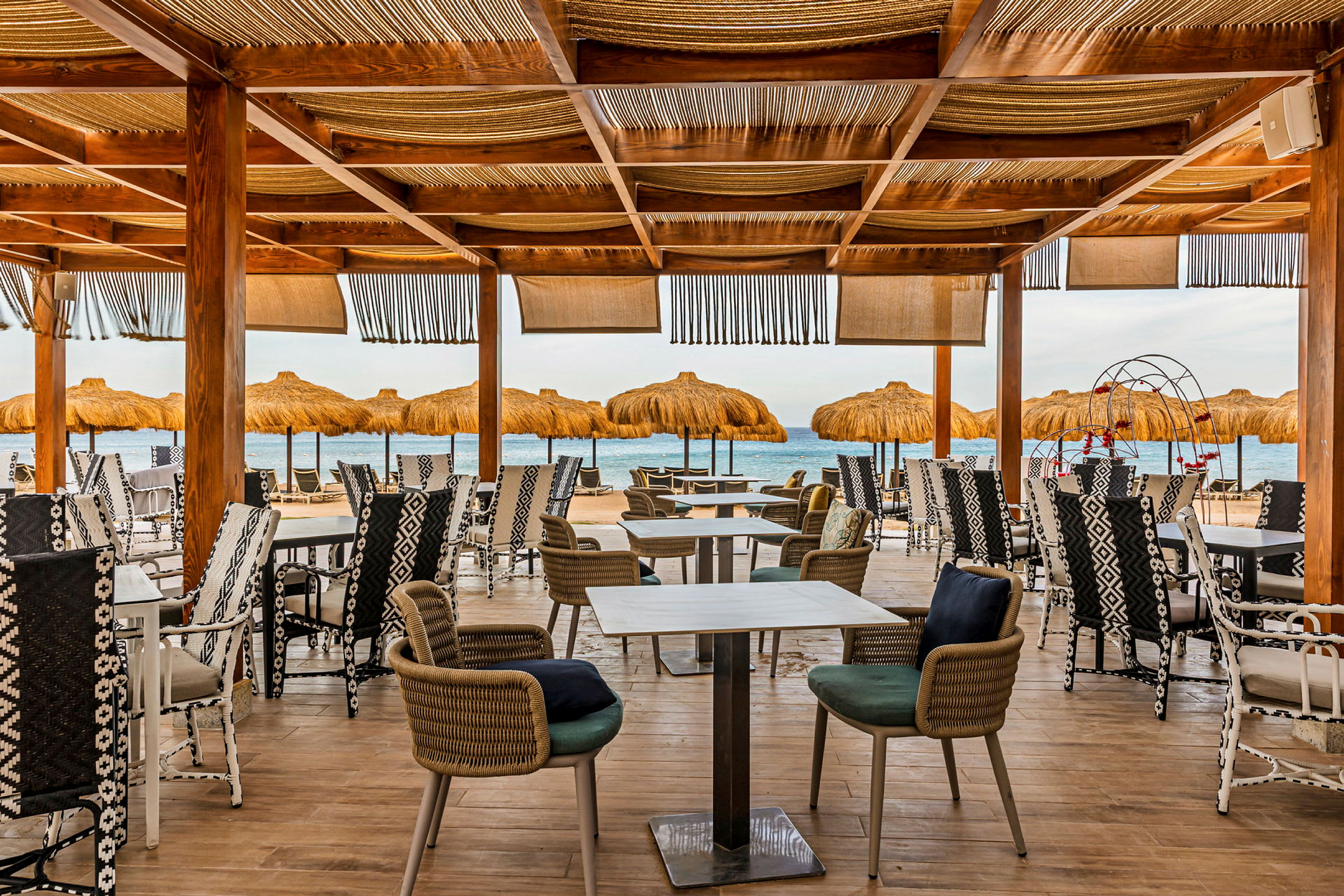 En utendørs restaurant med trestolper og stoler, med utsikt over havet og parasoller på stranden. (AI-generert tekst)