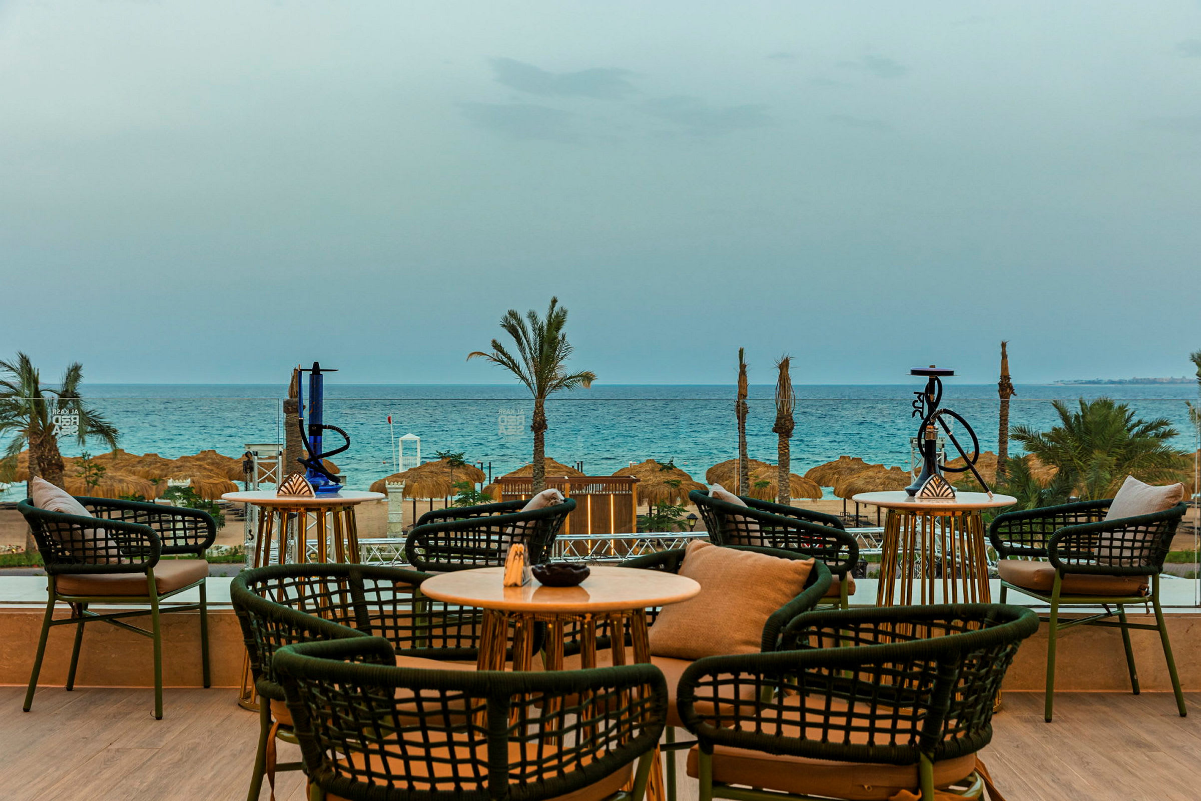 En utsikt fra en terrasse med sittemøbler, bord og vannpiper som vender mot stranden og havet i bakgrunnen. (AI-generert tekst)