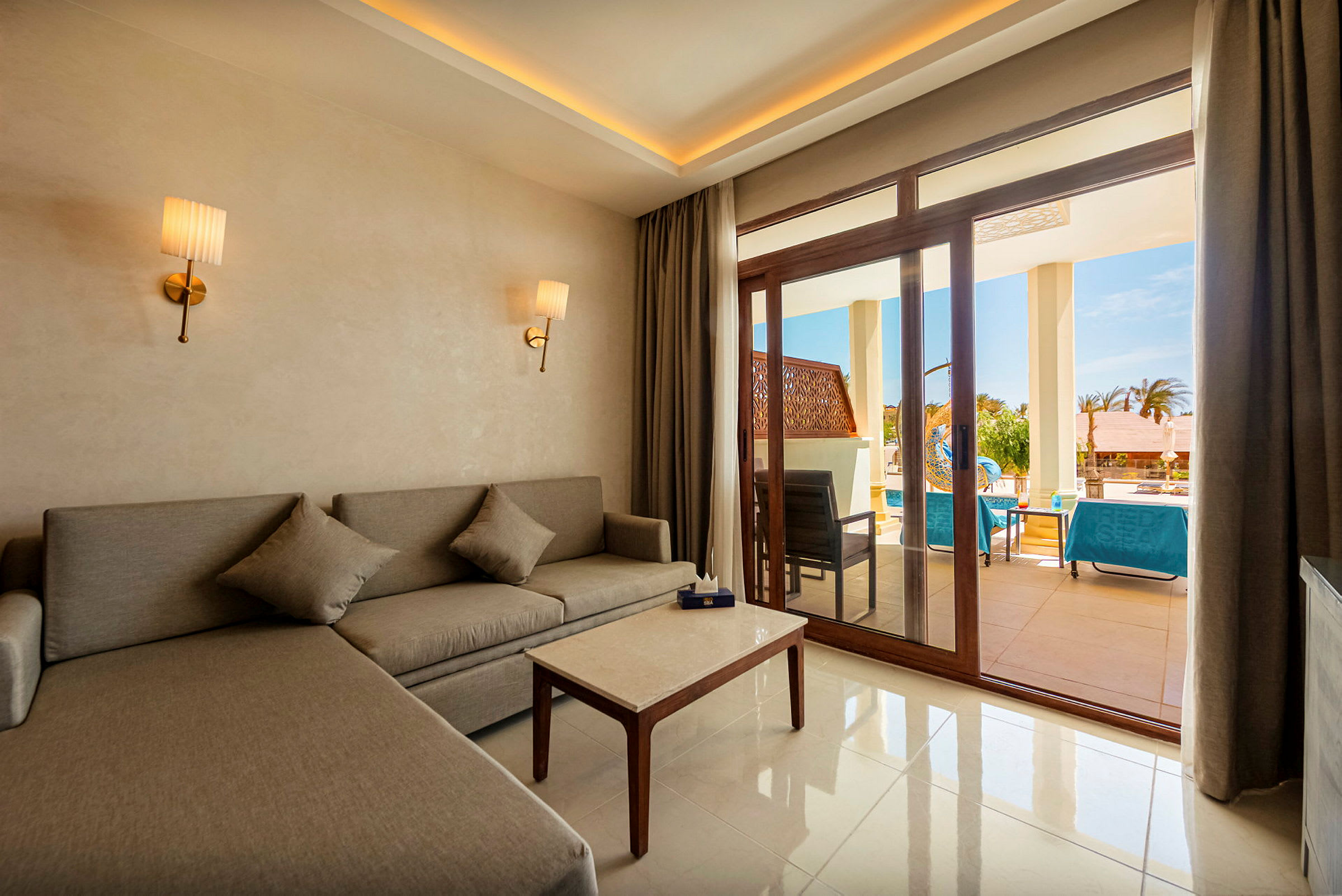 En hotellsuite med en grå sofa, et sofabord og to vegglamper. Utenfor glassdørene sees en terrasse med sittemøbler og solsenger ved et basseng. (AI-generert tekst)