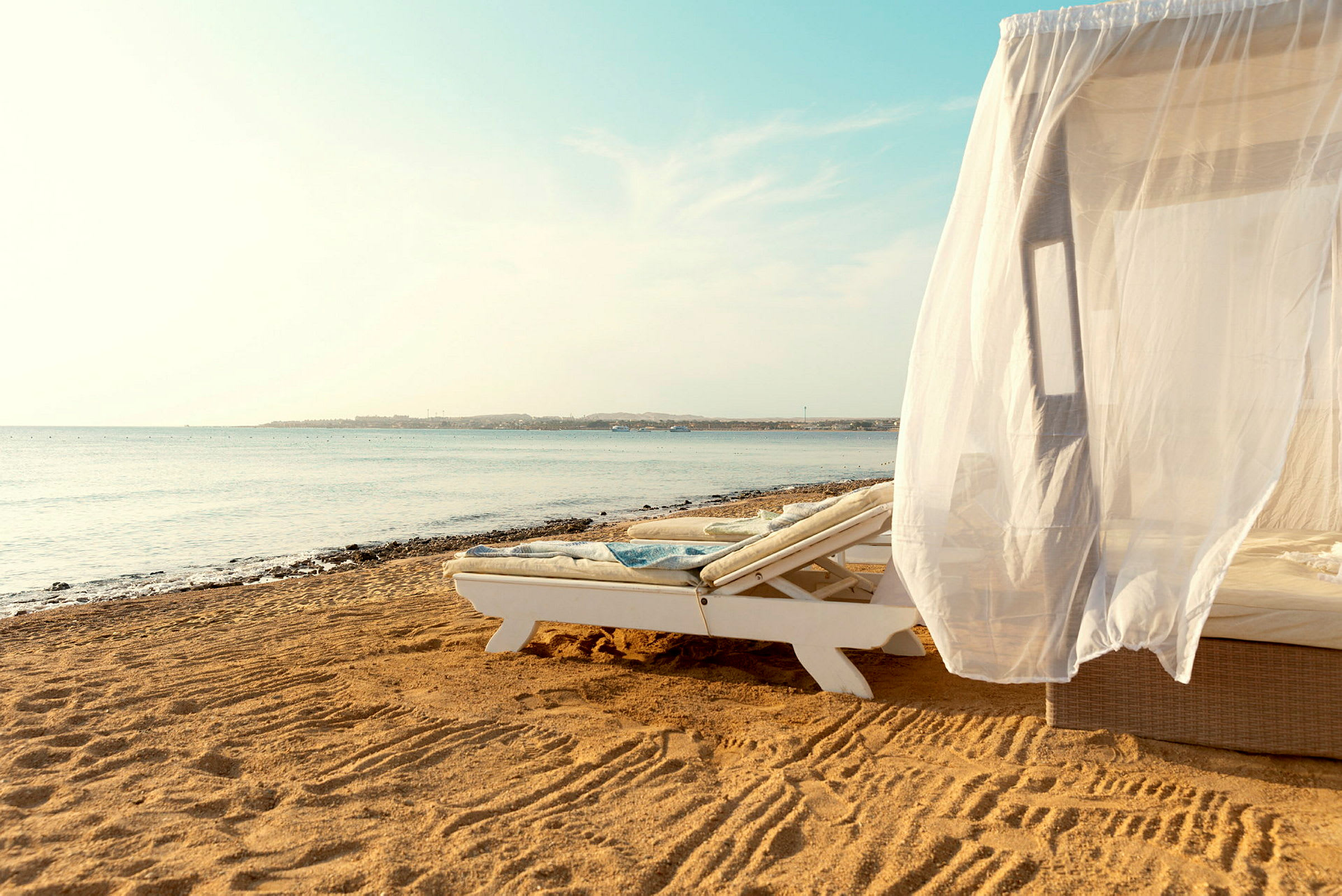 En solseng med et hvitt draperi på en sandstrand ved havet. (AI-generert tekst)