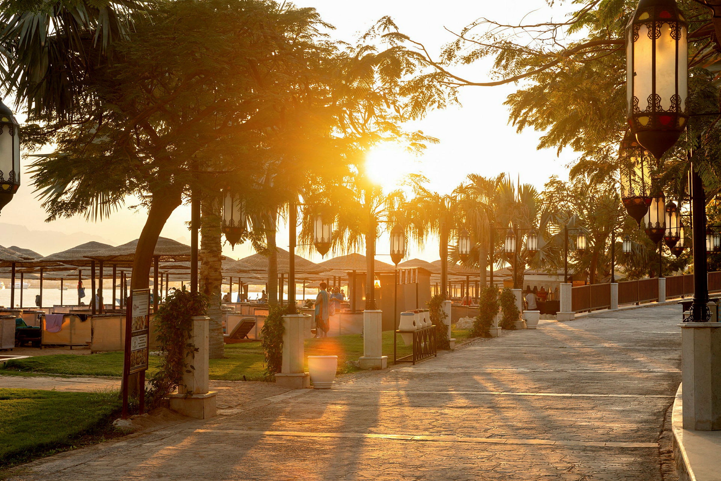 Solnedgang ved en strandpromenade med parasoller og bord. (AI-generert tekst)