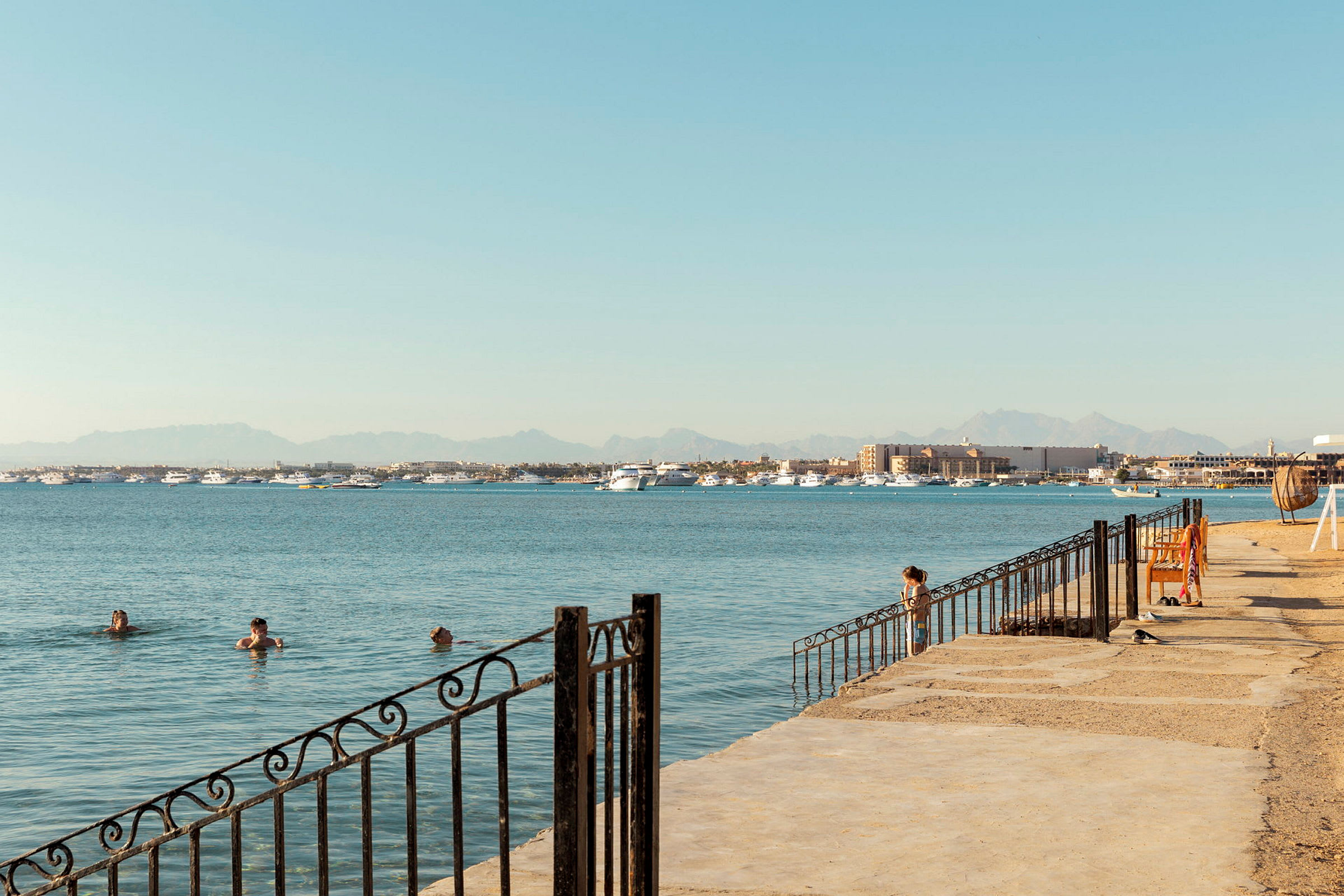 En strandpromenade med personer som bader i havet, båter i det fjerne og fjell i bakgrunnen. (AI-generert tekst)