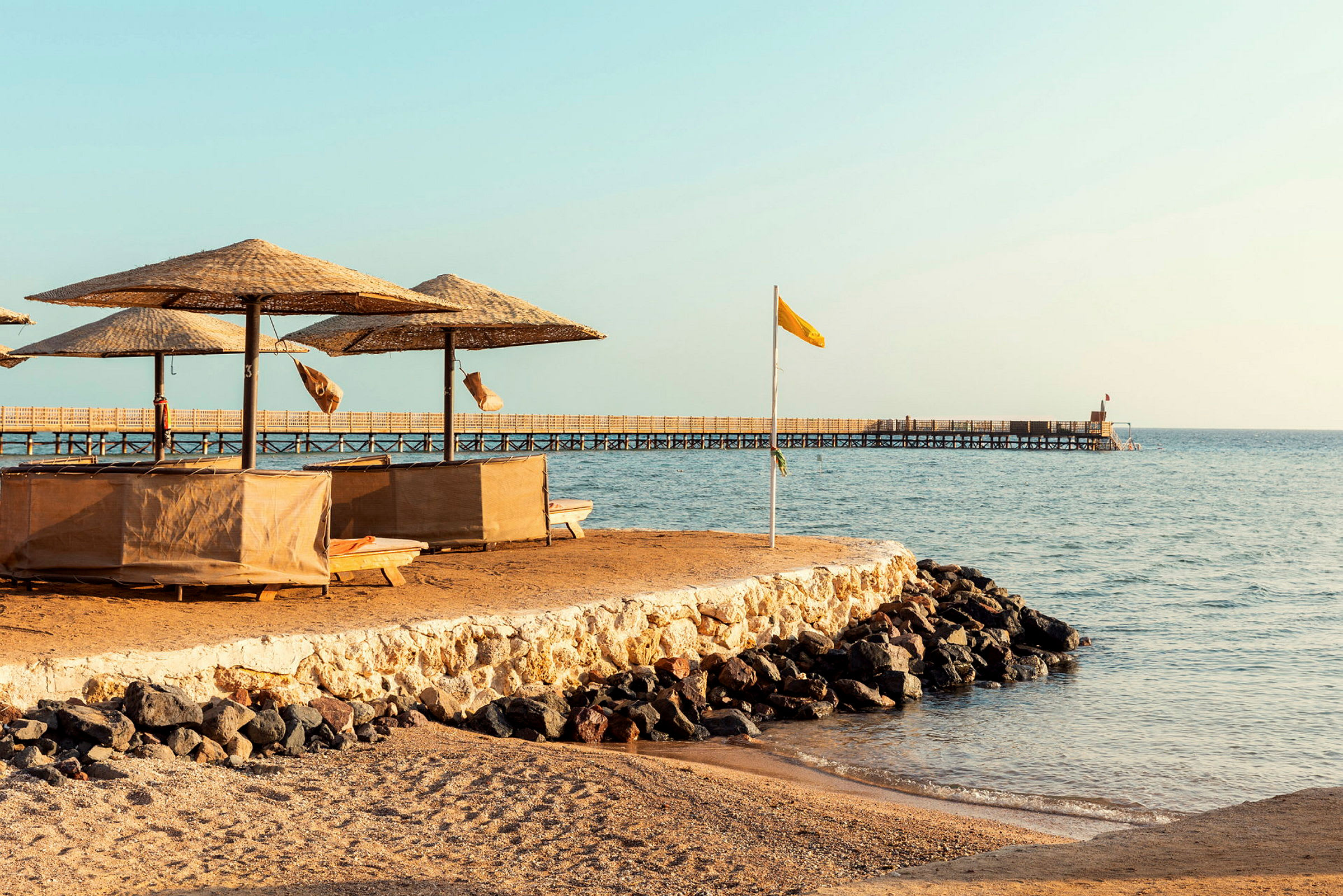 Strand med parasoller og solsenger, samt en brygge som strekker seg ut i havet. (AI-generert tekst)