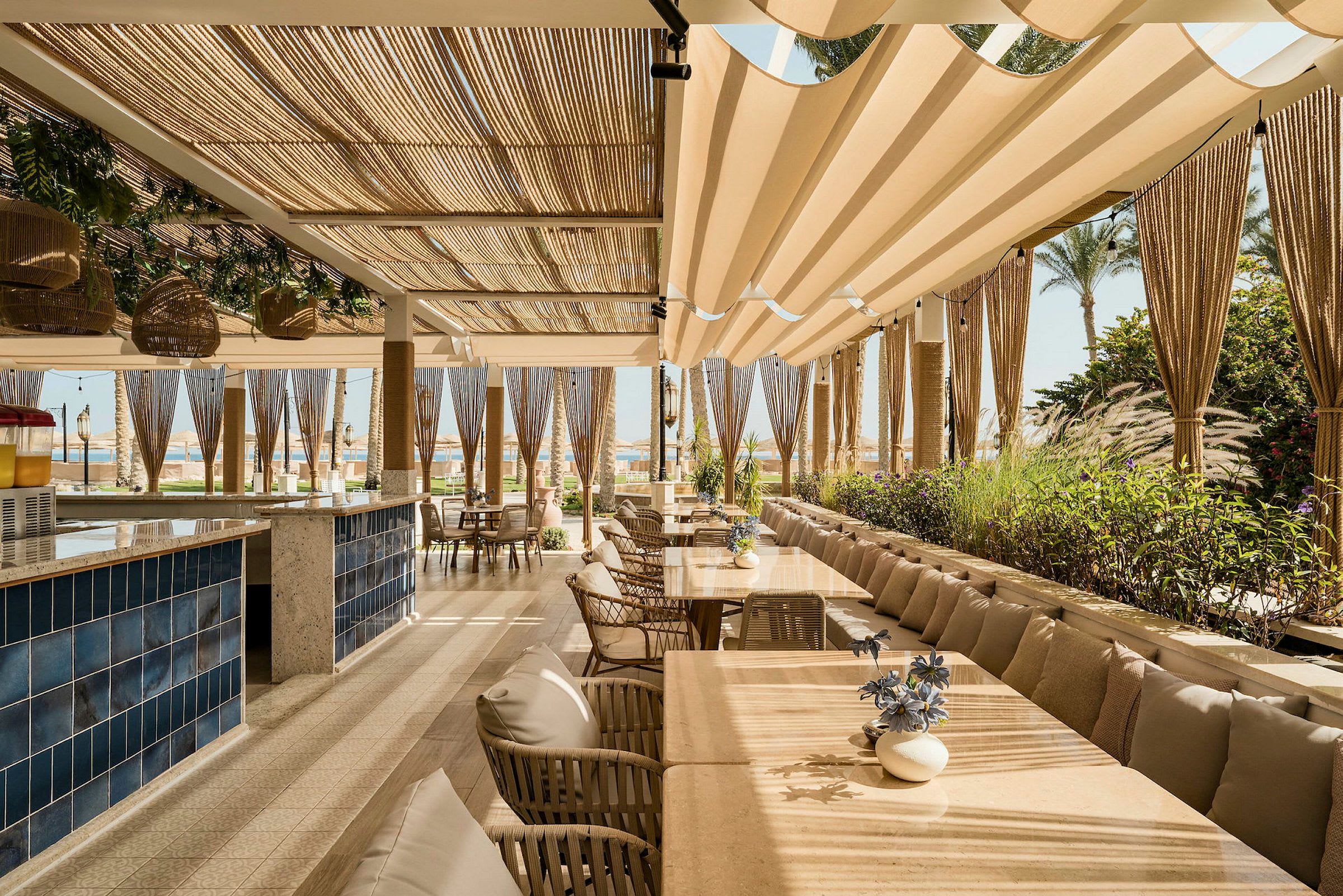 En restaurantterrasse med bord, stoler og beige solskjerming, omgitt av grønne planter. (AI-generert tekst)