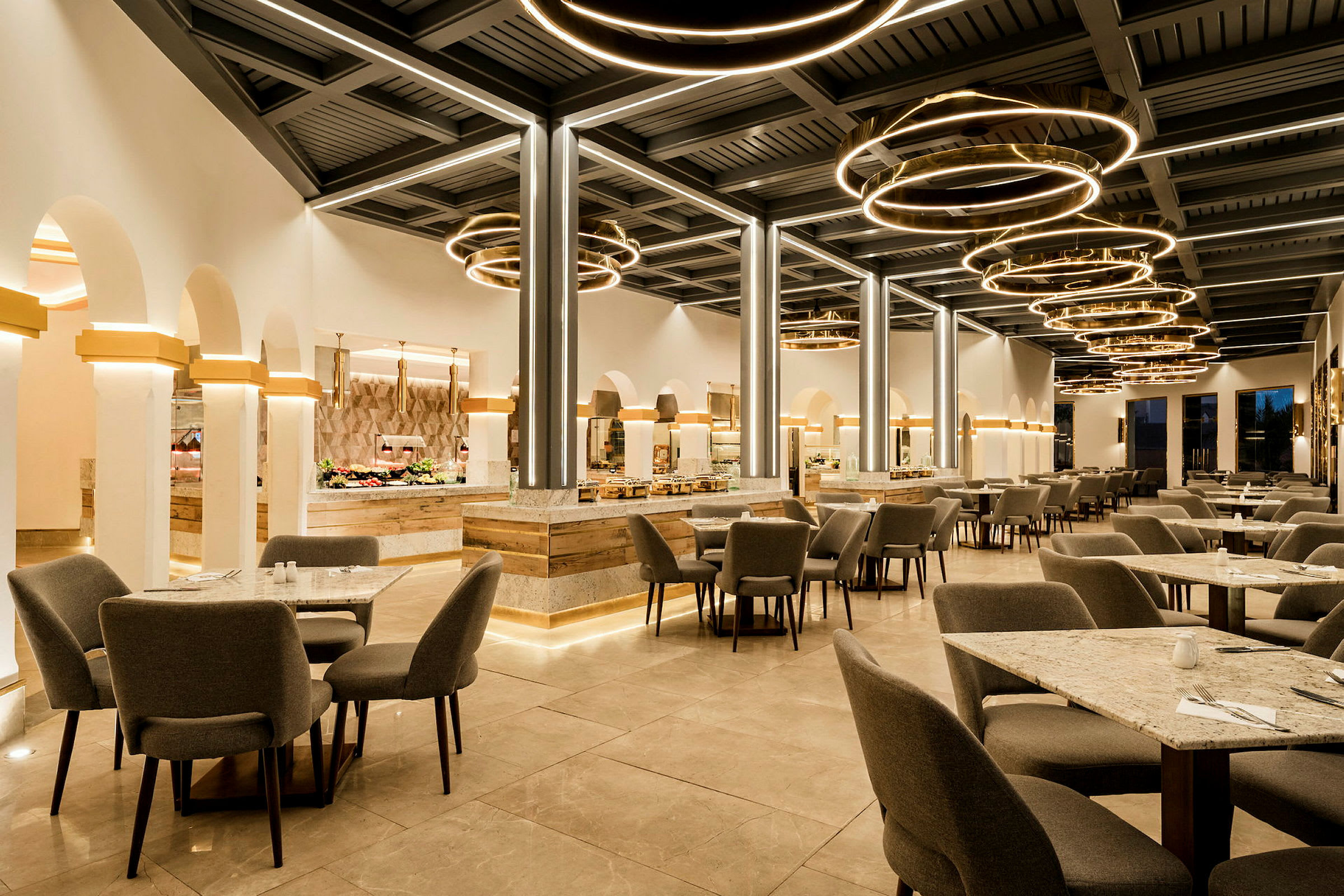 En moderne restaurant med grå stoler og bord på et steingulv, lysende sirkelformede lamper henger fra taket. (AI-generert tekst)