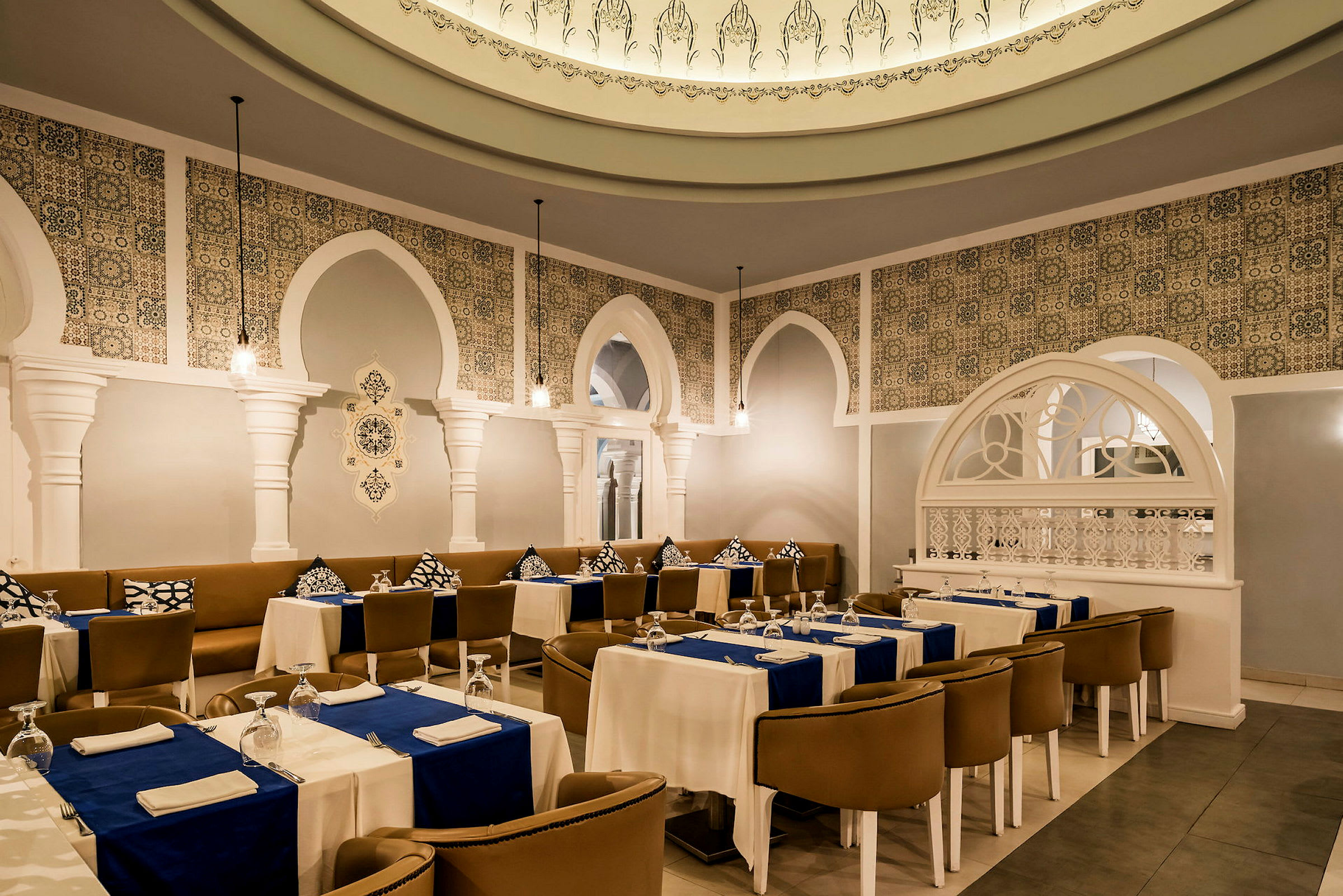 En elegant restaurant med hvite søyler, dekorativt flisemønster og oppdekkede bord med blå duker. (AI-generert tekst)