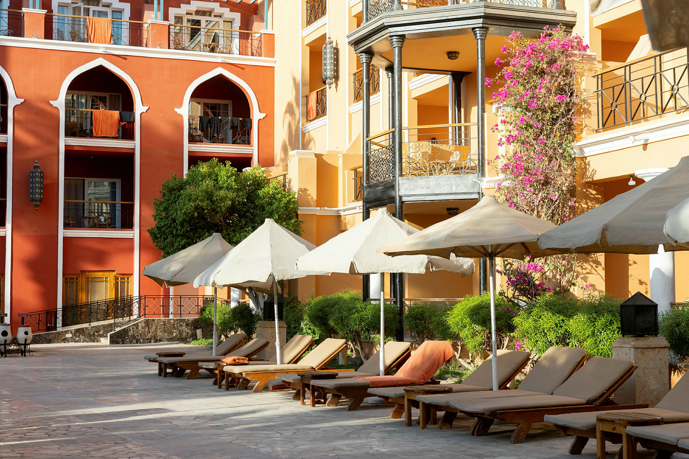 En hotellbygning med terrasser, parasoller og solsenger ved en steinlagt uteplass. (AI-generert tekst)