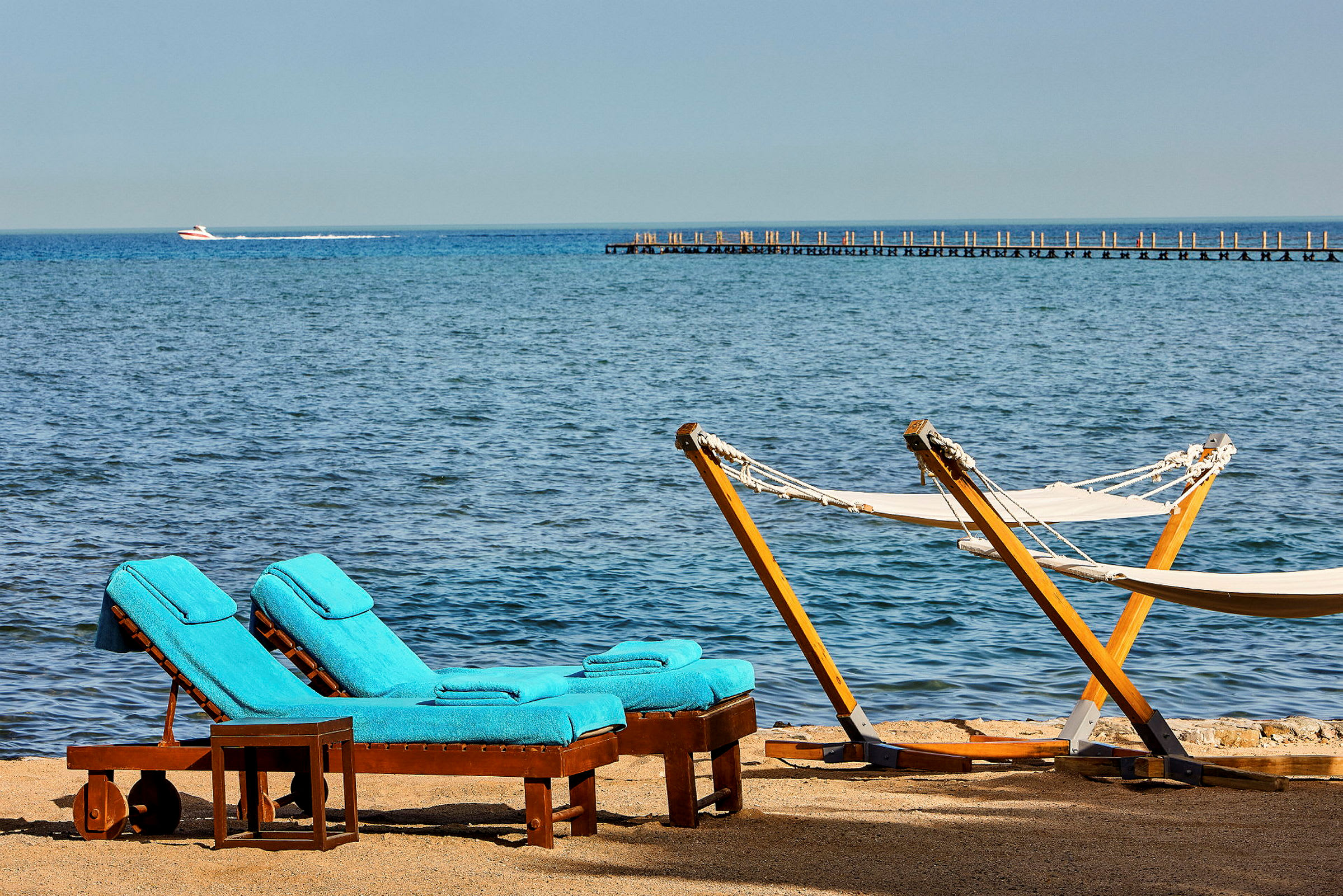To solsenger med blå puter og to hengekøyer ved en strand med havutsikt. (AI-generert tekst)