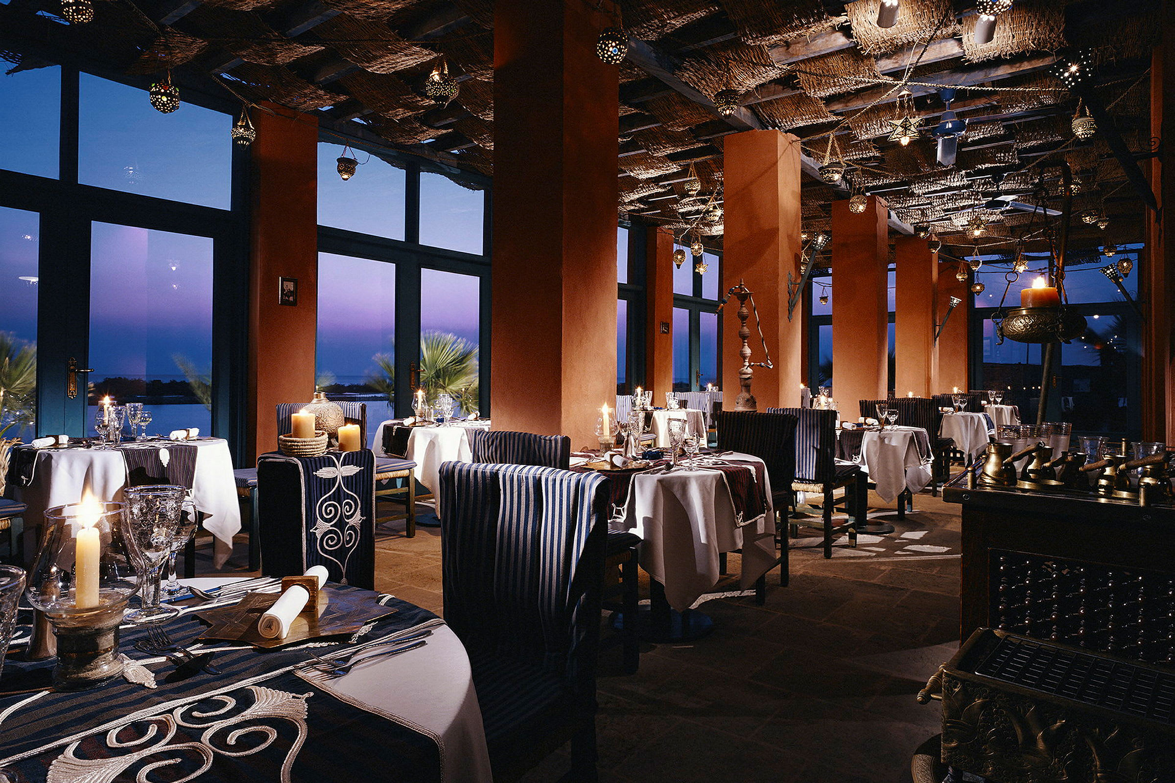 Restaurant med belysning, bord og stoler, samt store vinduer mot skumringen. (AI-generert tekst)