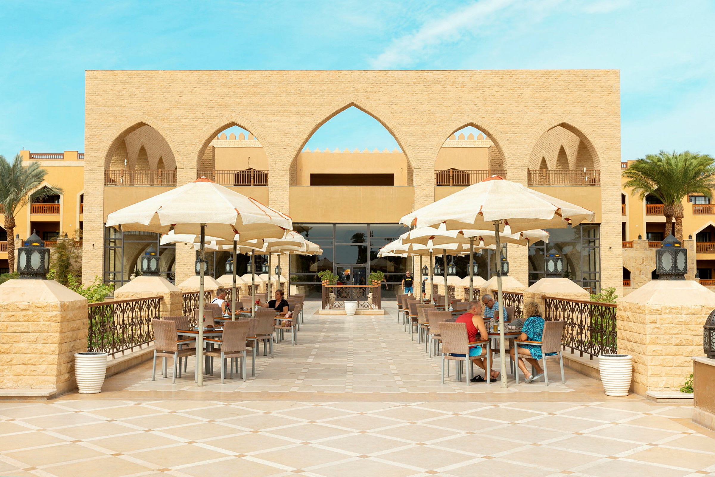 Restaurantterrasse med parasoller ved hotell, bygning i bakgrunnen. (AI-generert tekst)