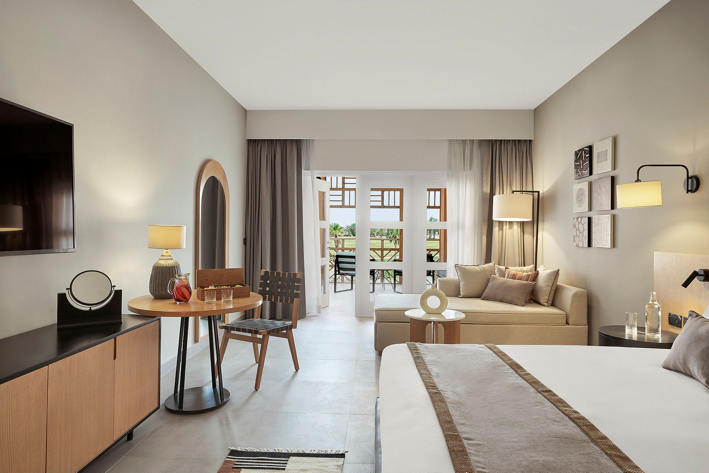 Et moderne hotellrom med grå og beige fargetoner, inkludert en seng, sittegruppe, bord og en balkong. (AI-generert tekst)