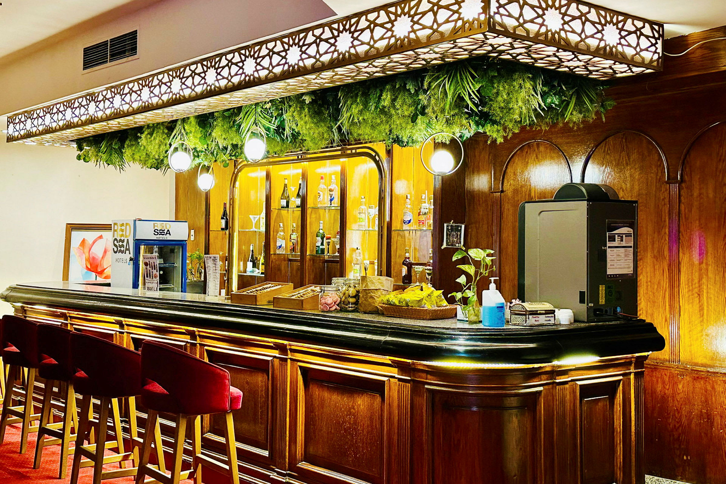 En elegant bar med røde barstoler, en trebenk og flasker på en bakgrunn med belysning. Planter og dekorasjoner henger ovenfor. (AI-generert tekst)