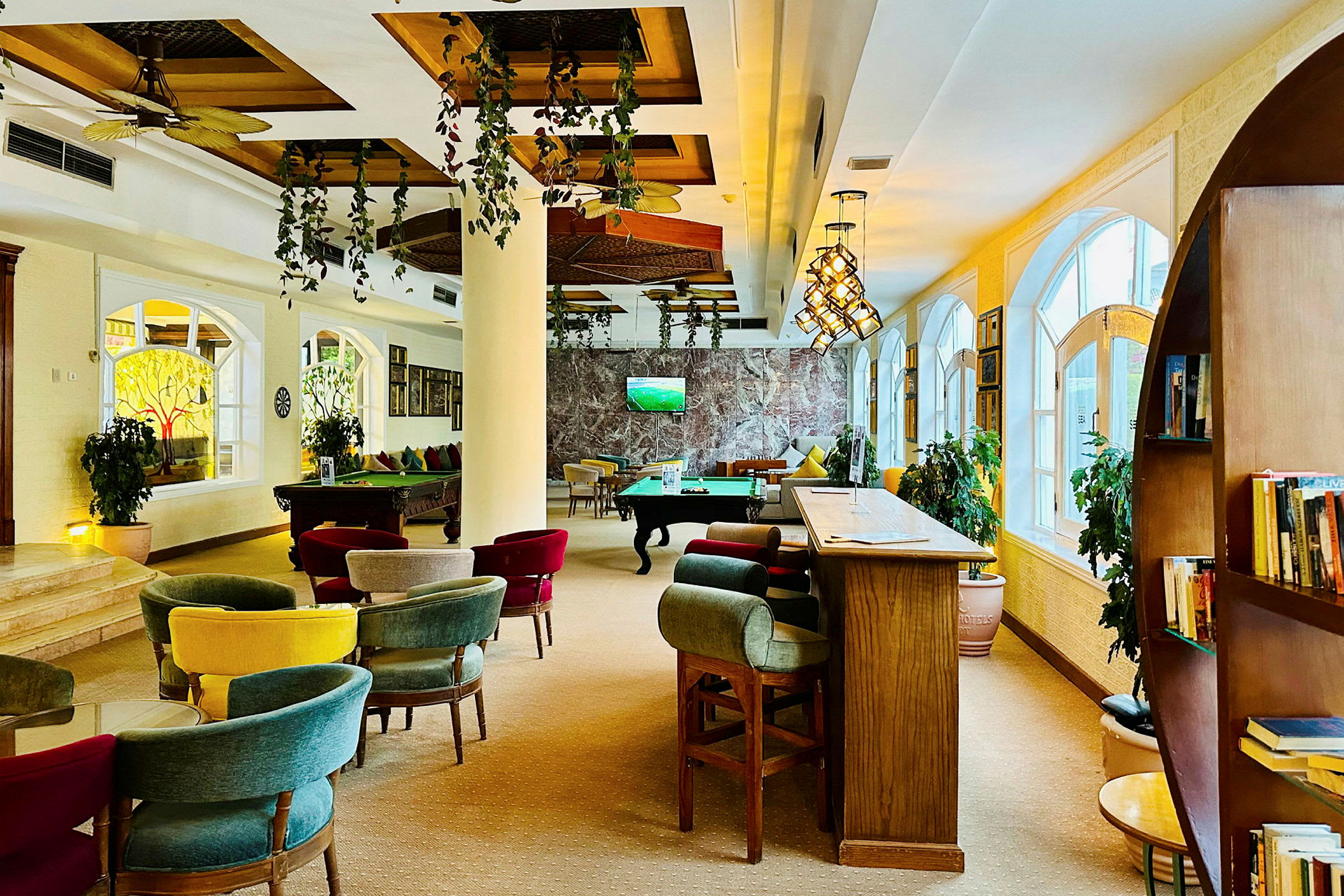 Et innendørsområde med sitteplasser, biljardbord og en bar med høye sitteplasser. Planter og store vinduer dekorerer rommet. (AI-generert tekst)
