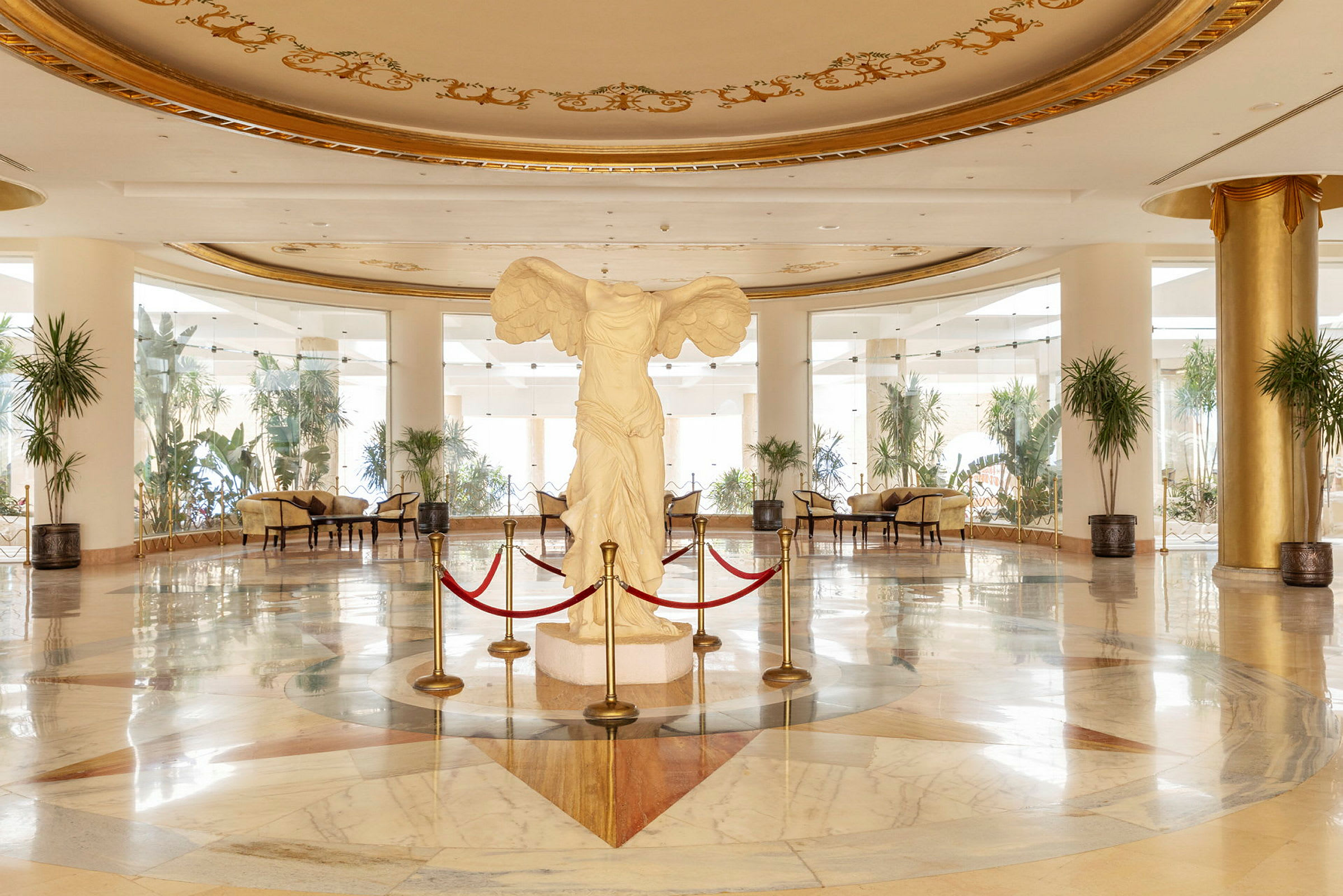 Interiør av en elegant hotellobby med en hvit statue omgitt av røde tau i sentrum. (AI-generert tekst)