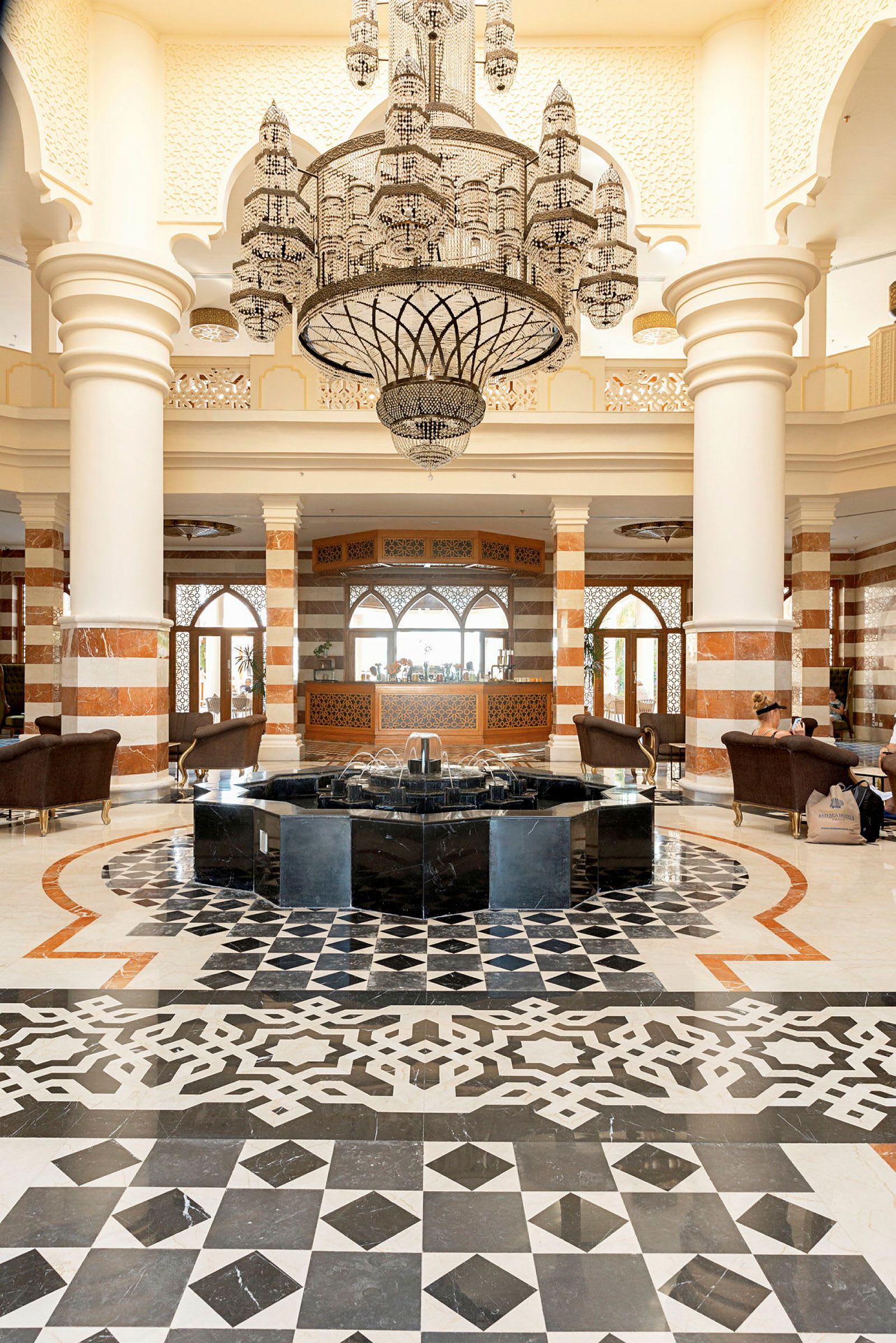 Elegant hotellobby med mønstret gulvbelegg, fontene i midten og en stor lysekrone som henger over. (AI-generert tekst)
