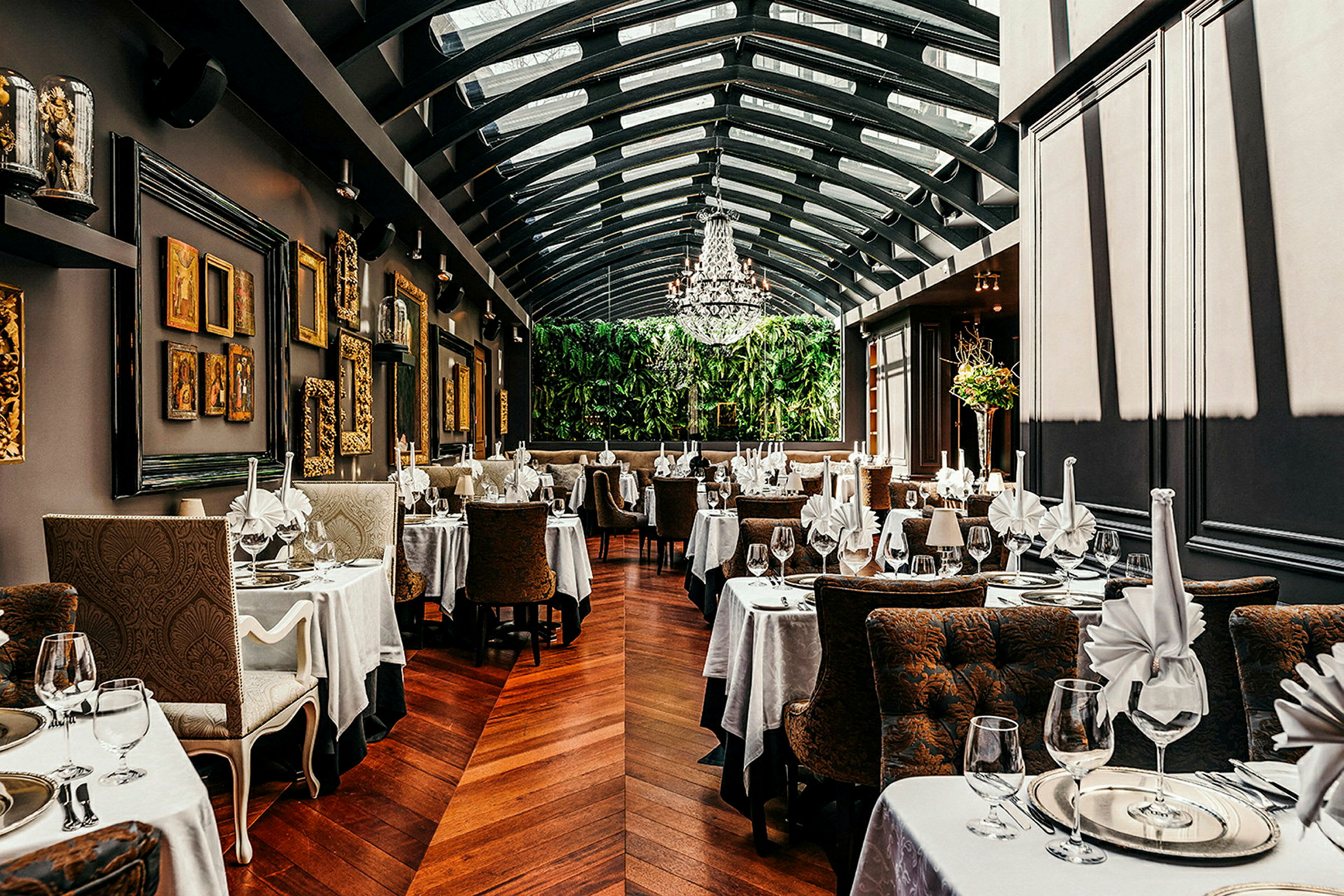 Restaurant med elegante bord, duker og glass. Veggene er dekorert med kunst, og rommet har glasstak. (AI-generert tekst)