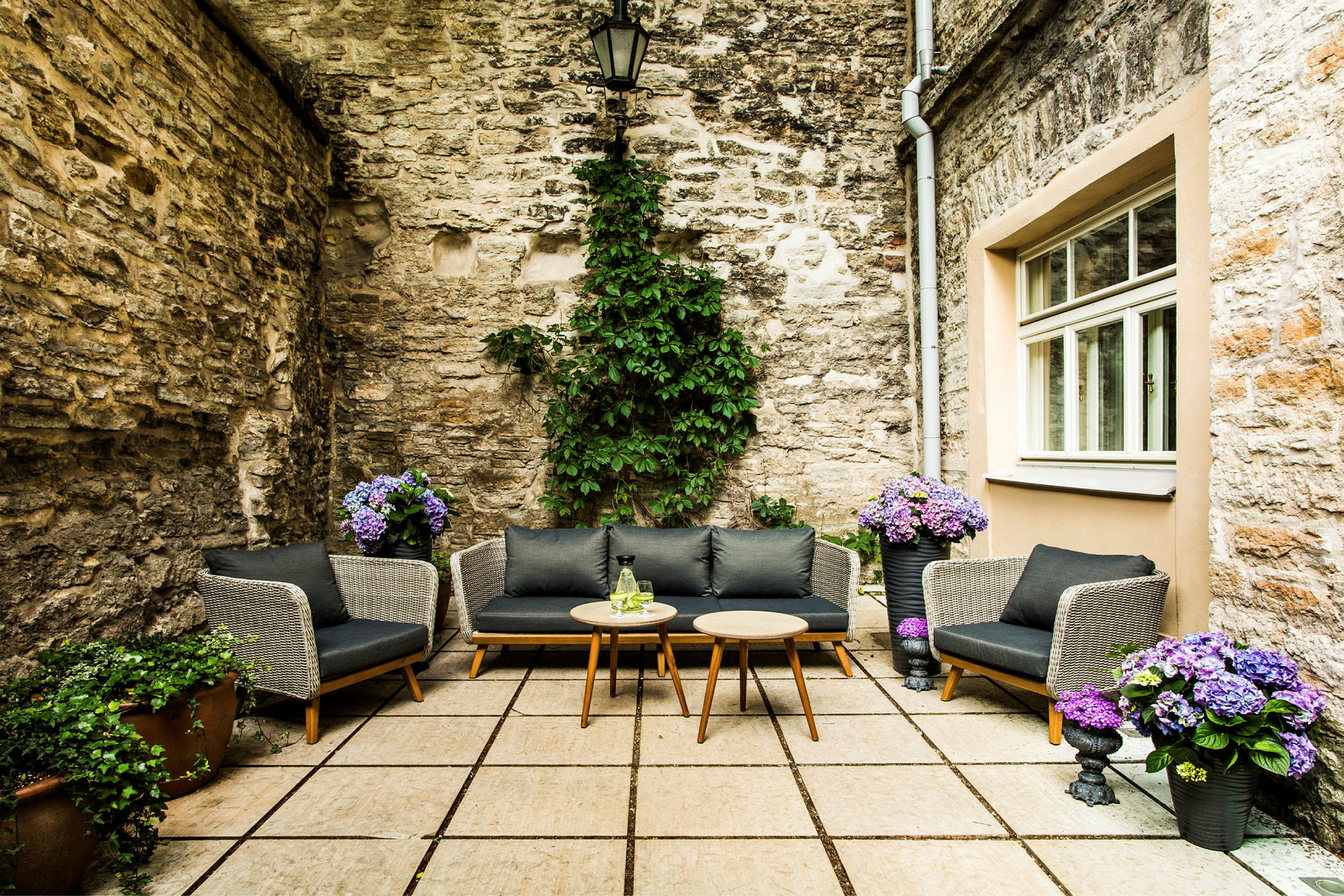 En indre gårdsplass med steinlagt gulv, steinvegger og koselige sitteplasser med puter. Potter med blomster og grønne planter pynter området. (AI-generert tekst)