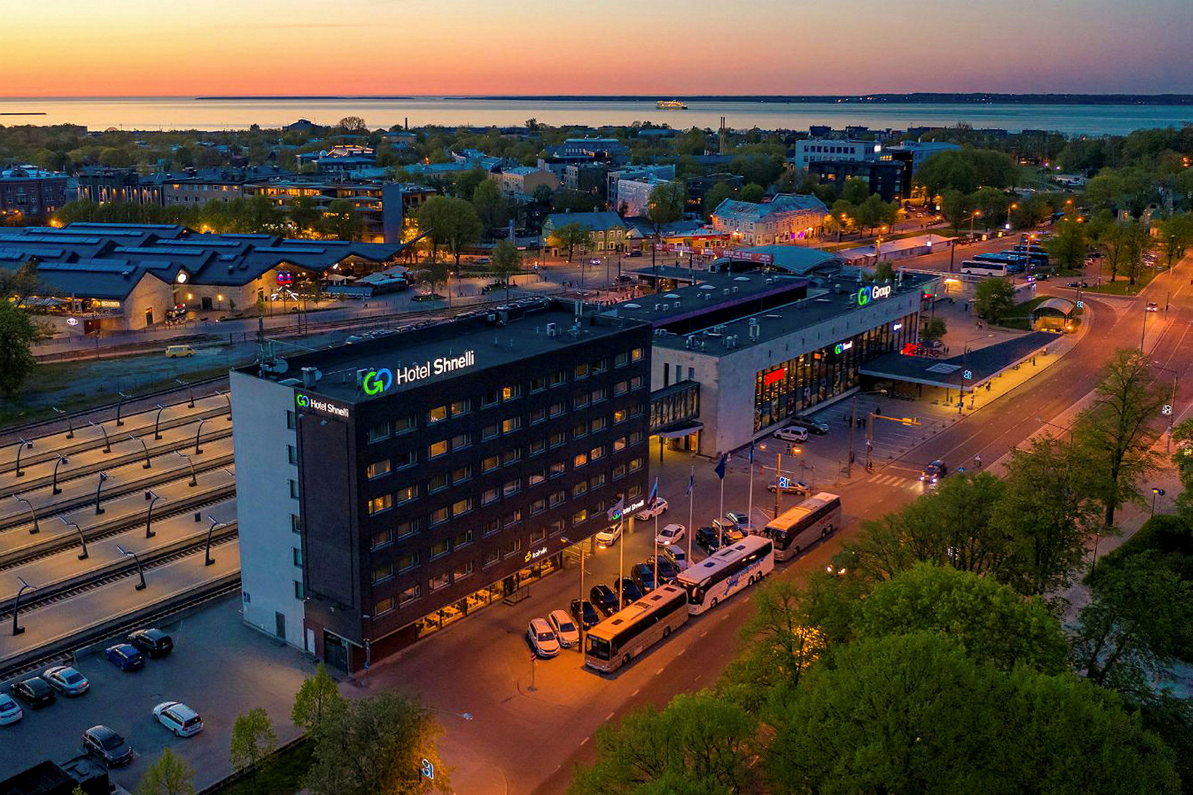 En større bygning med skiltet "Hotel Shnelli" er synlig ved en vei omringet av busser og biler. Området er opplyst på kvelden med en bakgrunn av trær og en havn. (AI-generert tekst)