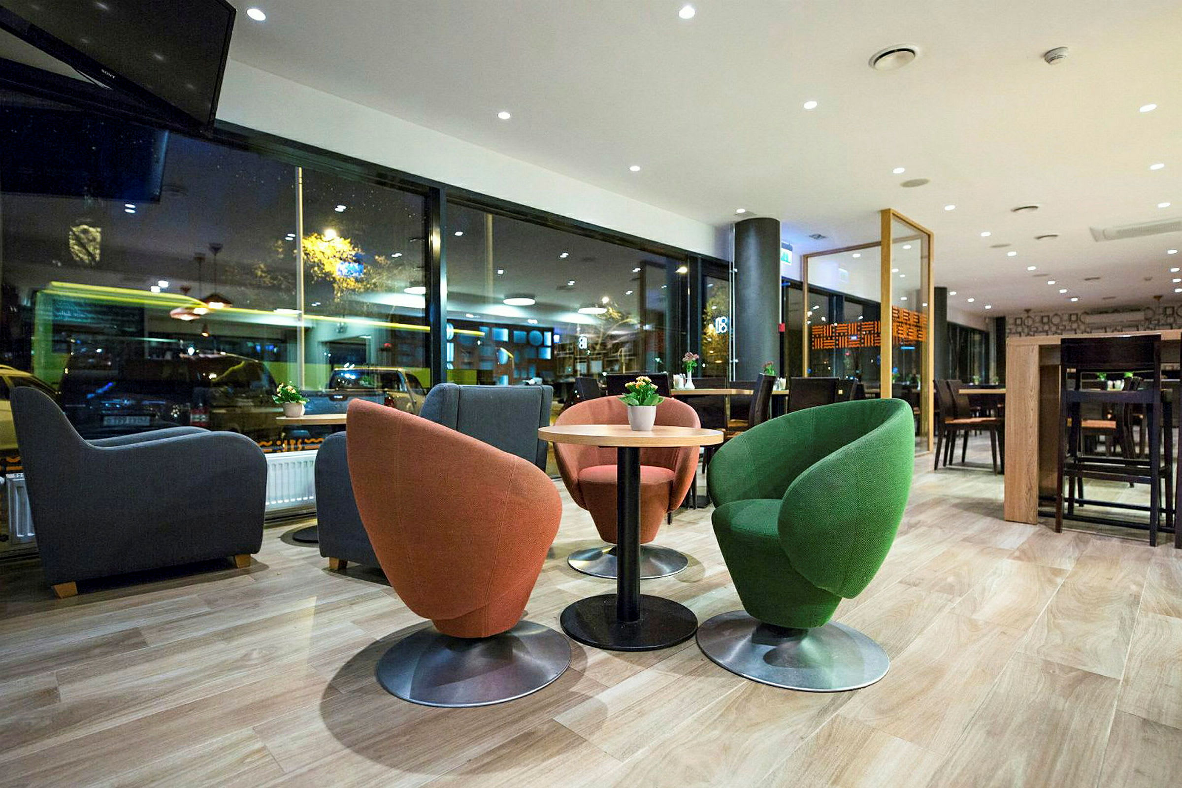 Et moderne kafé eller restaurant med fargerike stoler rundt et bord, store glassvinduer og kveldslys utenfor. (AI-generert tekst)