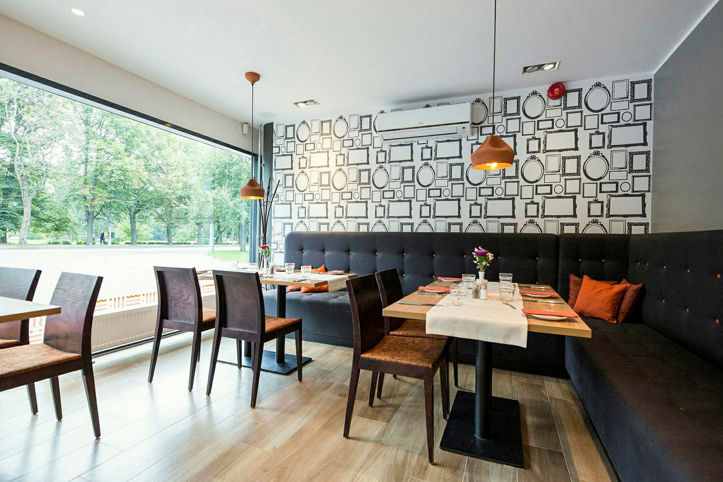 En moderne innredet kafé eller restaurant med store vinduer, trebord og stoler samt en lang sofa langs veggen. (AI-generert tekst)