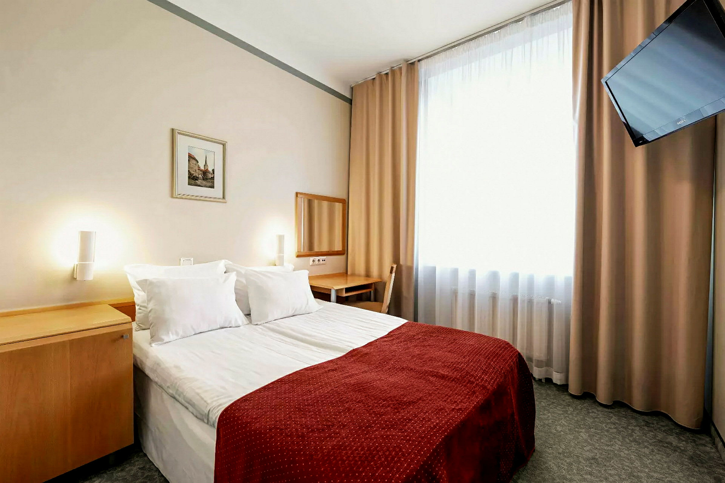 Et hotellrom med en dobbeltseng, rødfarget sengeteppe. Et speil og skrivebord står ved siden av veggen. Et vindu med lyse gardiner og en veggmontert TV finnes i rommet. (AI-generert tekst)