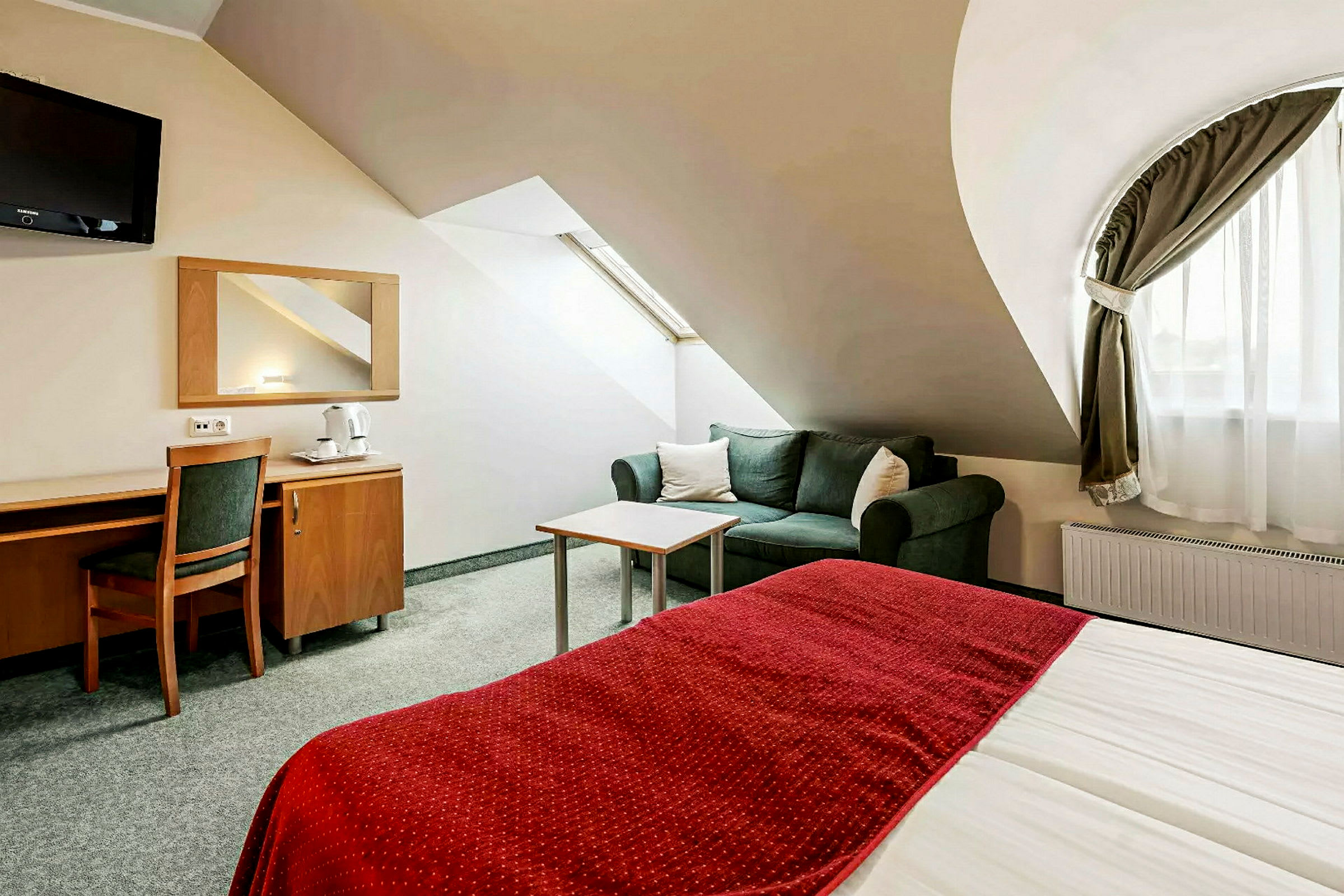 Et hotellrom med en seng med rødt teppe, en sofa og et skrivebord med stol. (AI-generert tekst)