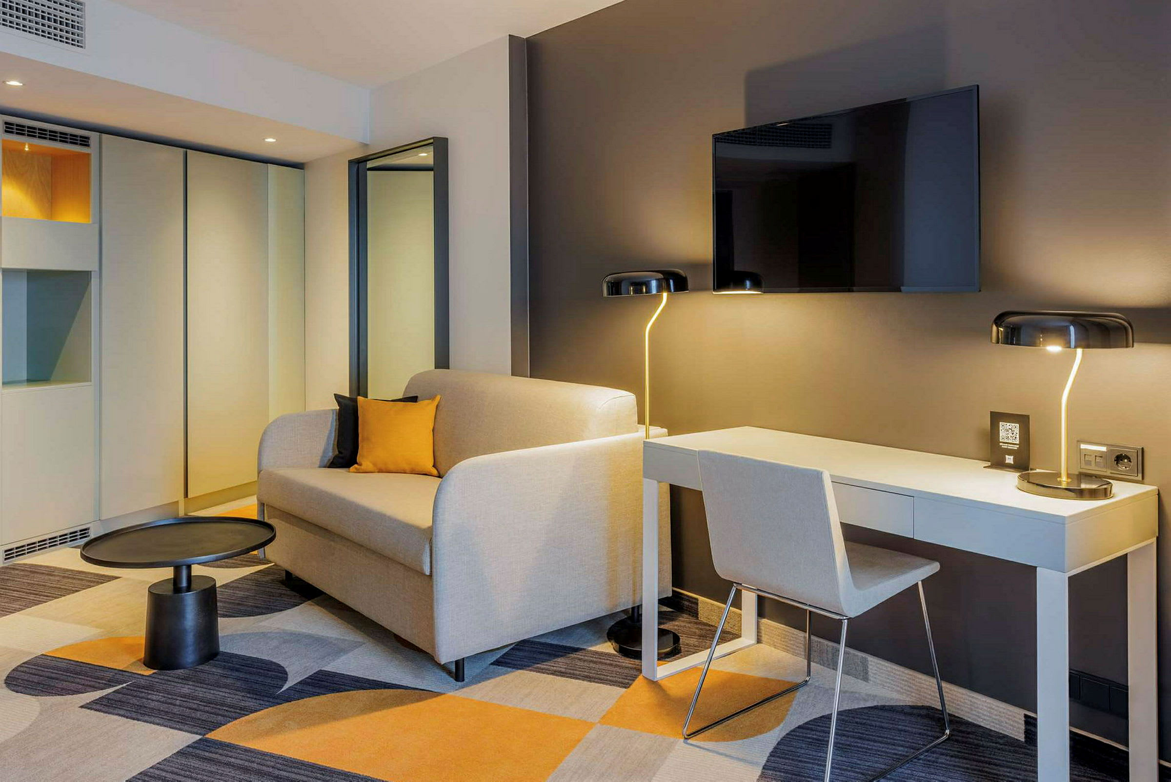 Et moderne hotellrom med en sofa, et skrivebord og en veggmontert TV. (AI-generert tekst)