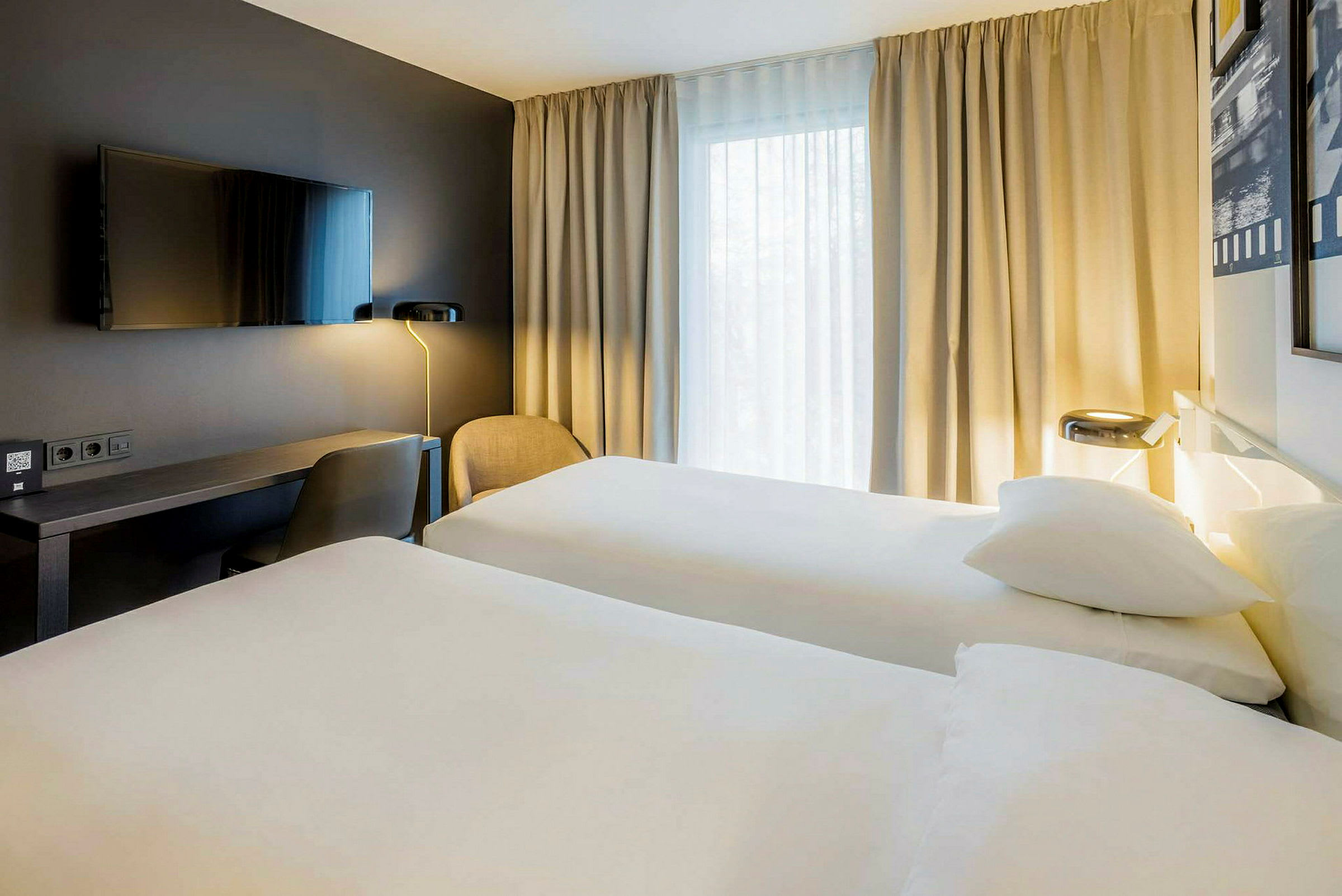 Hotellrom med to enkeltsenger, et skrivebord, en stol og en veggmontert TV, samt gardiner ved et vindu. (AI-generert tekst)