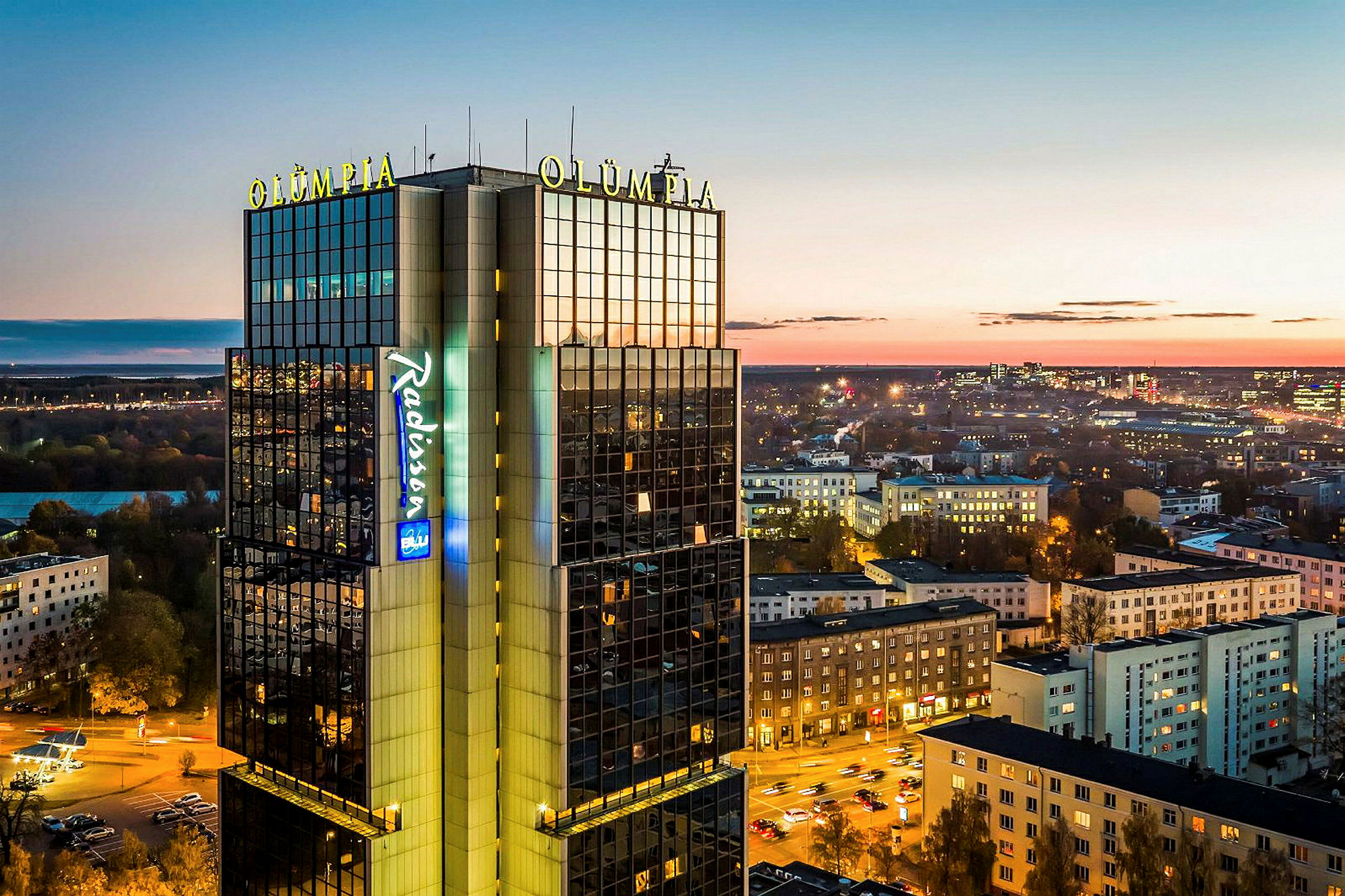 En høy bygning merket "Olümpia" og "Radisson" med byutsikt i skumringen. (AI-generert tekst)