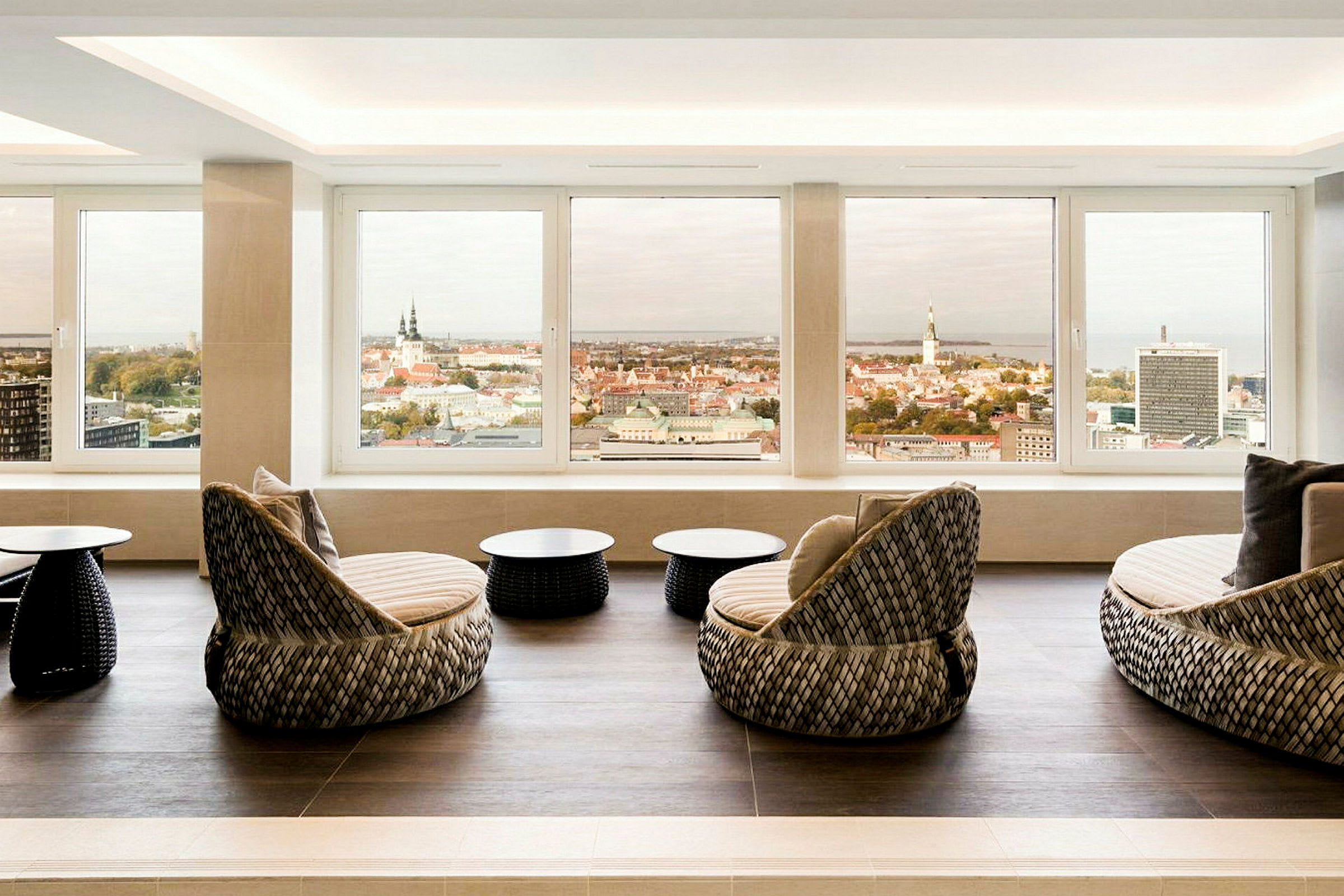 Et rom med store vinduer og utsikt over en bysilhuett. I forgrunnen er det runde sofaer og små bord. (AI-generert tekst)