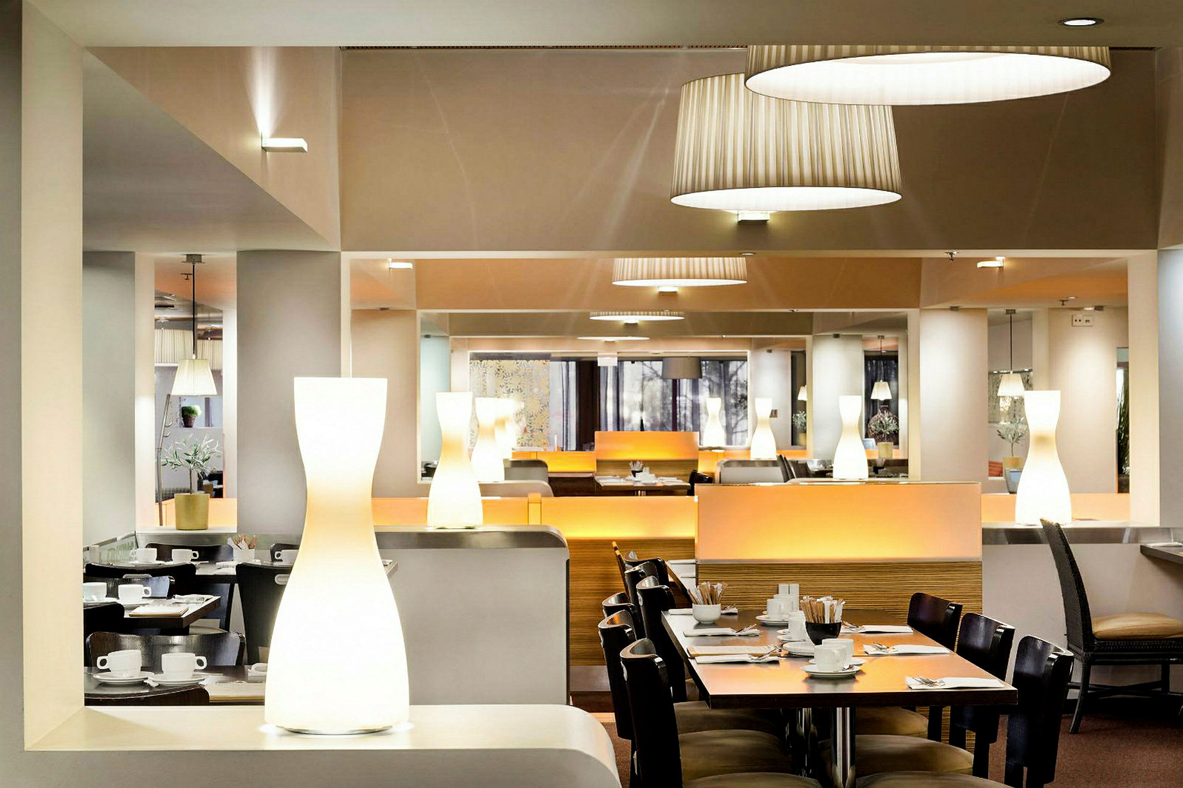 En elegant spisesal med moderne interiør, der bord er dekket med servise og bestikk. Belysningen består av tak- og bordlamper. (AI-generert tekst)
