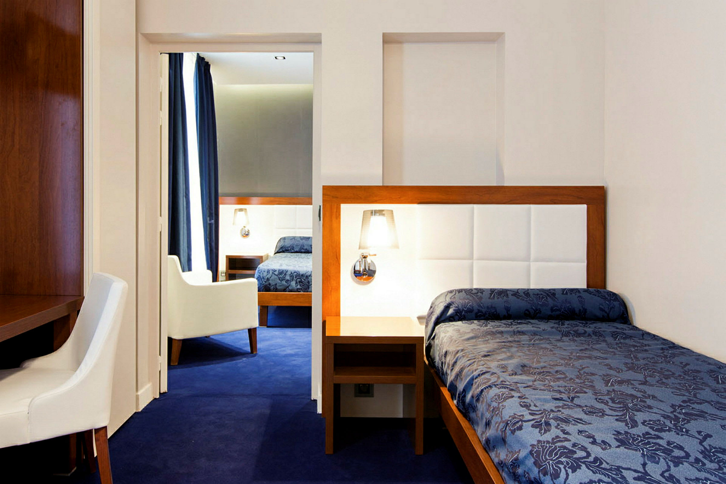 Et hotellrom med blått teppe, en enkeltseng med blått sengeteppe og lyse møbler. (AI-generert tekst)