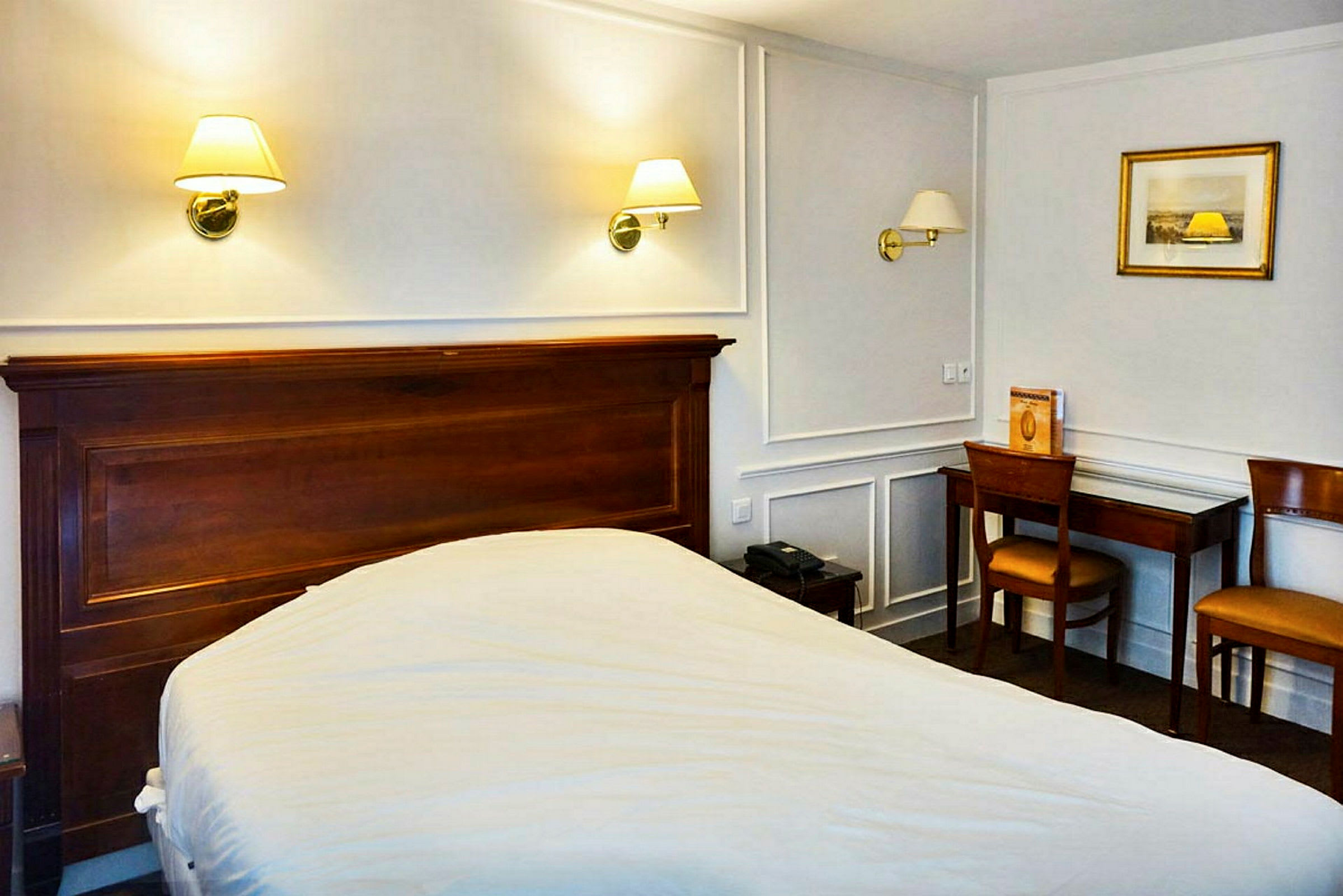 Et hotellrom med en seng, trebord og stoler samt vegglamper over sengen. (AI-generert tekst)