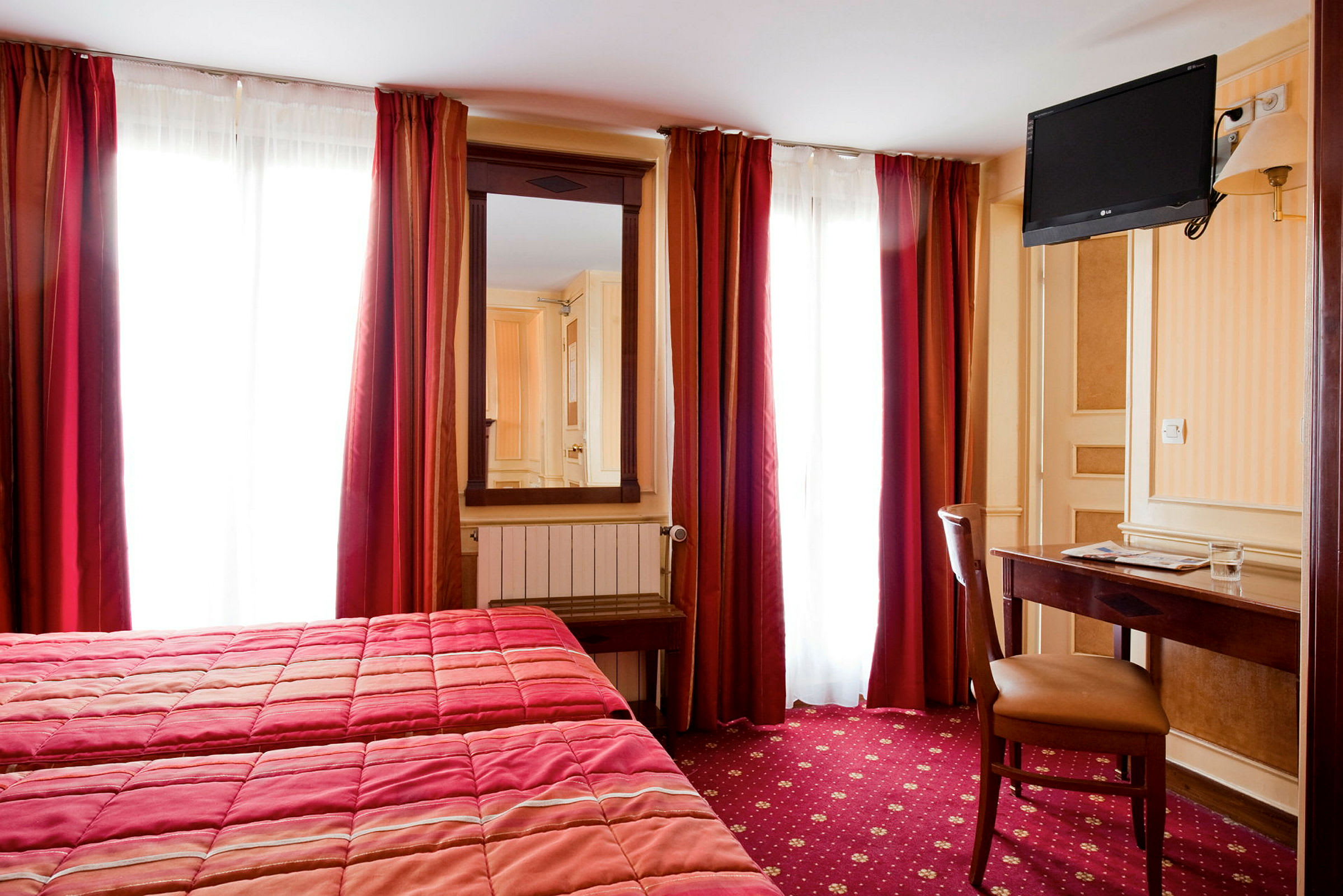 Et hotellrom med to senger, et speil, en veggmontert TV og et skrivebord med stol. (AI-generert tekst)