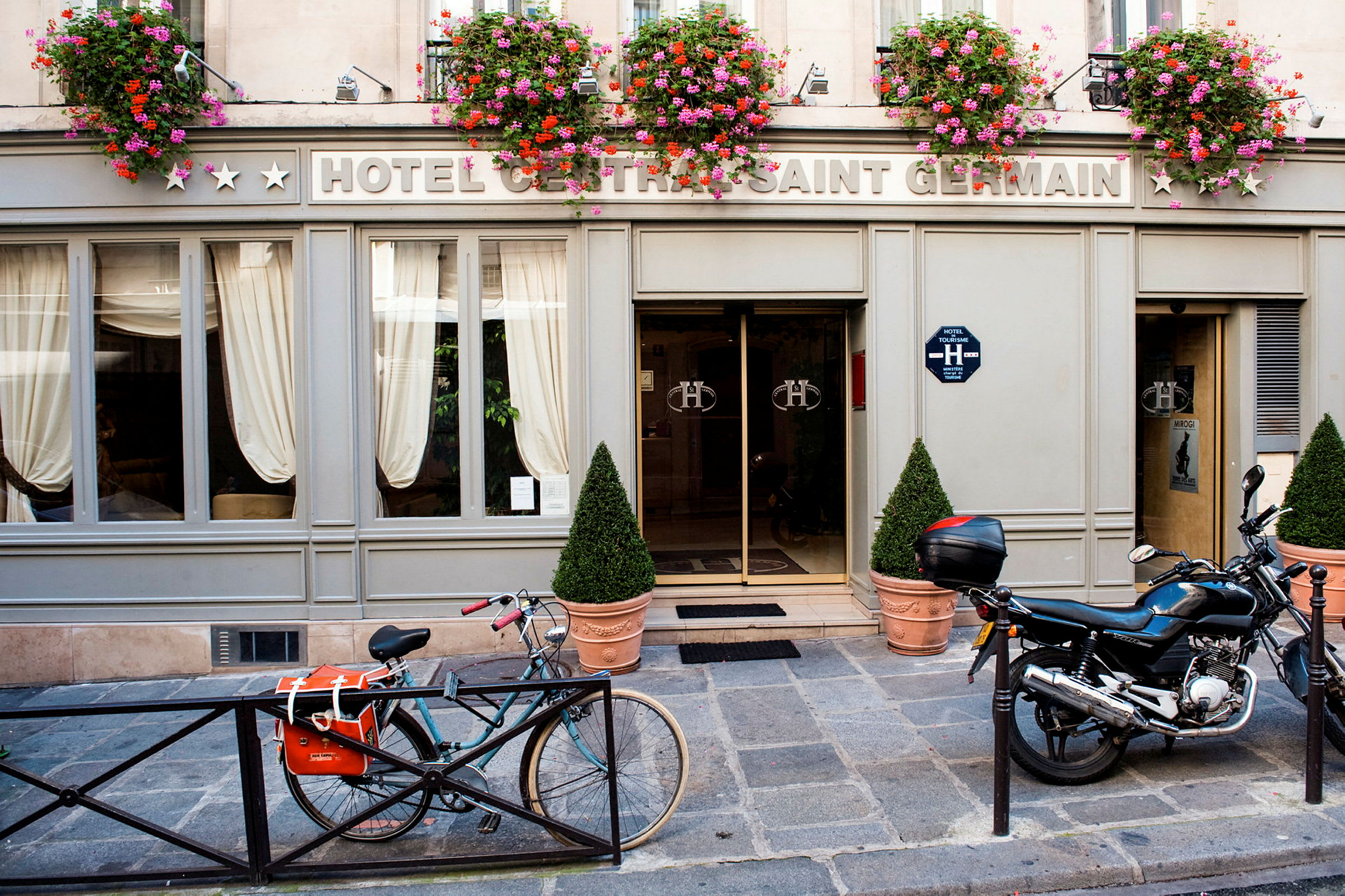Eksteriør av et hotell med skiltet 'Hotel Central Saint Germain' med blomsterpotter over inngangen samt en sykkel og motorsykkel parkert foran. (AI-generert tekst)