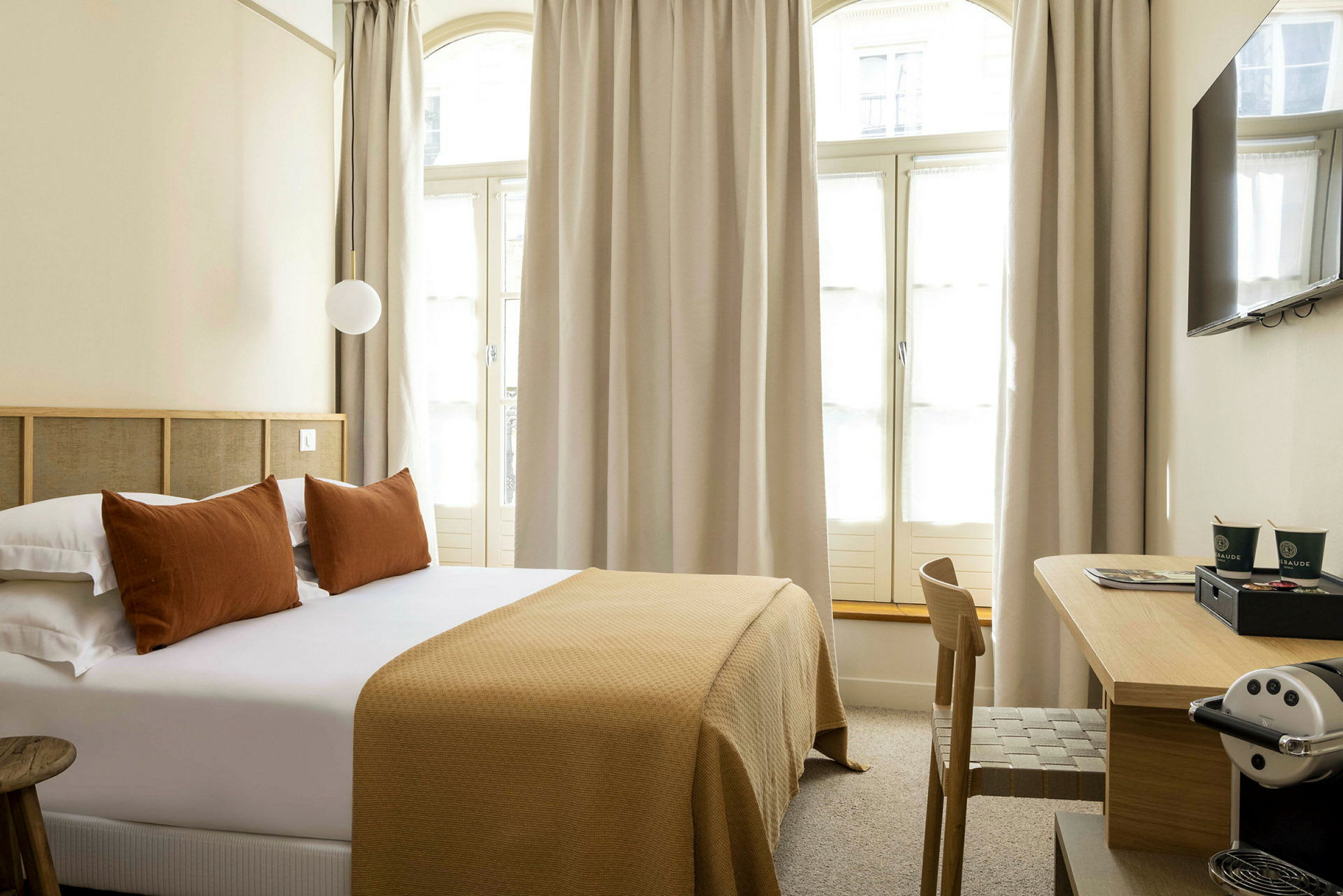 Hotellrom med en dobbeltseng, puter og et sengeteppe i brune toner samt et skrivebord med en kaffemaskin. (AI-generert tekst)