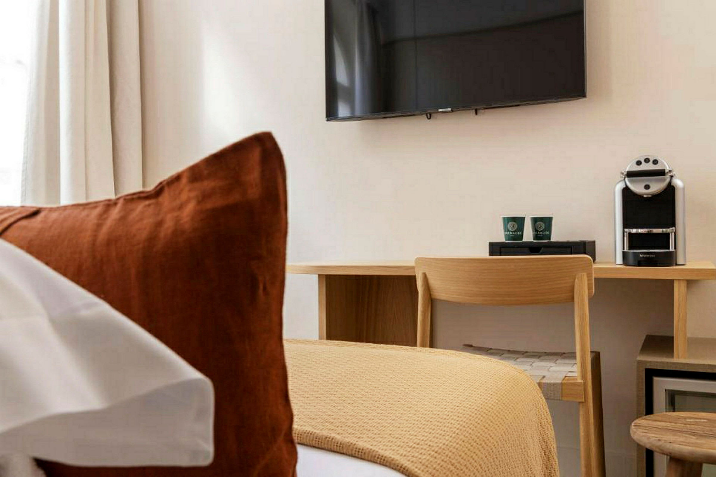 Et hotellrom med en seng i forgrunnen, en skrivebordplass med stol, kaffemaskin og en veggmontert TV. (AI-generert tekst)