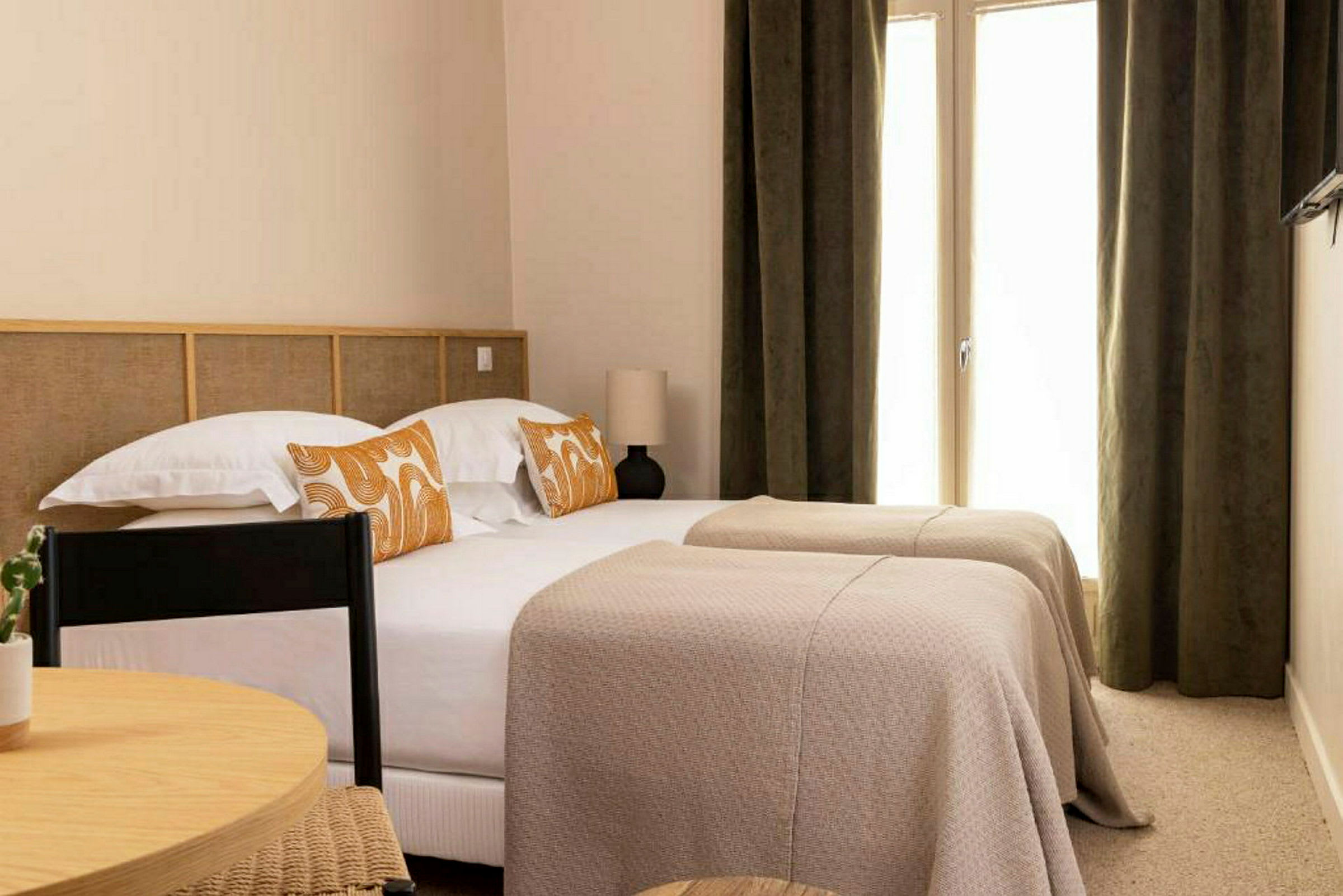 Et hotellrom med to enkeltsenger, dekket med beige sengetepper og dekorert med oransjemønstrede puter. (AI-generert tekst)