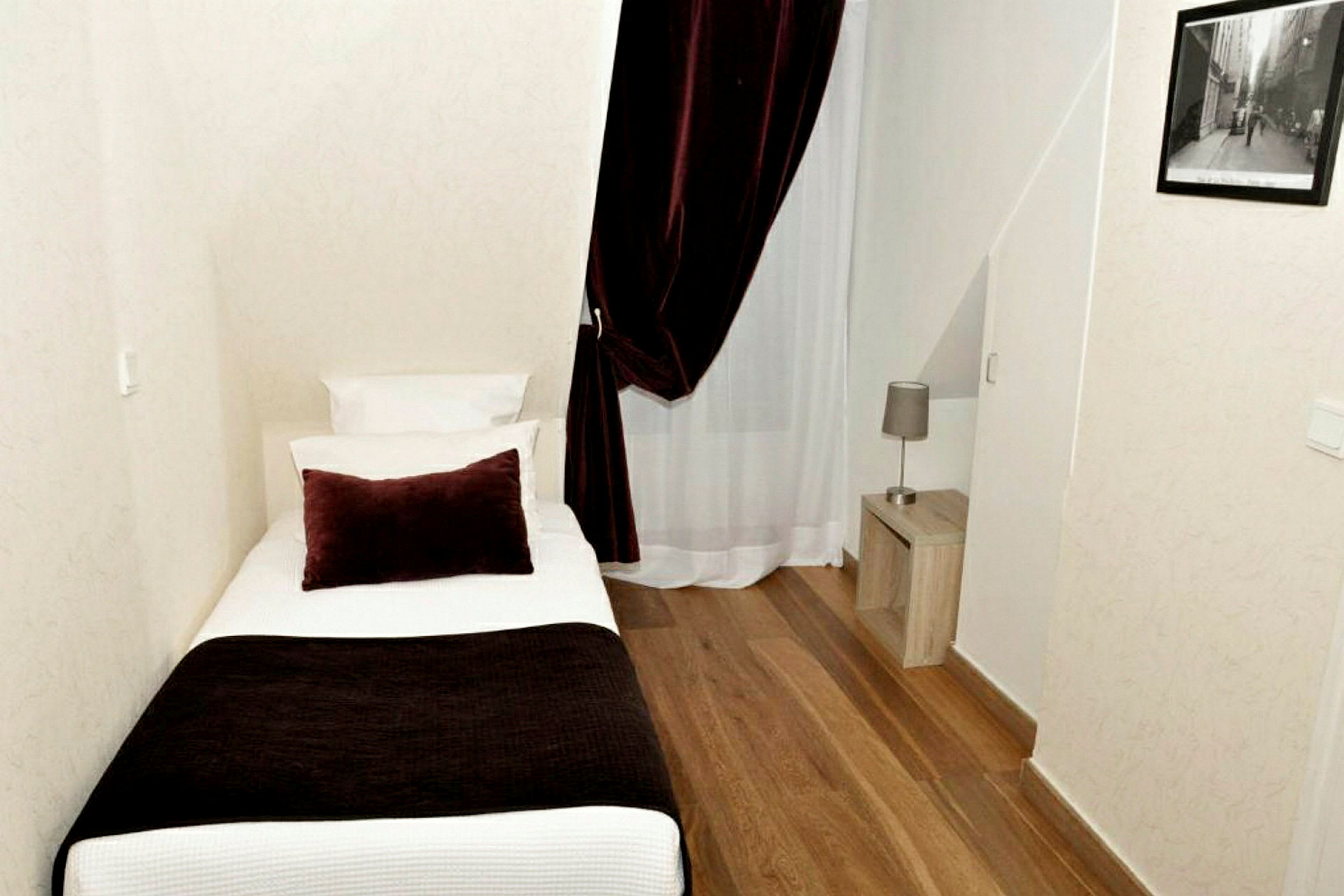 Et hotellrom med enkeltseng, nattbord og en lampe. Rommet har tregulv og et gardin foran et vindu. (AI-generert tekst)