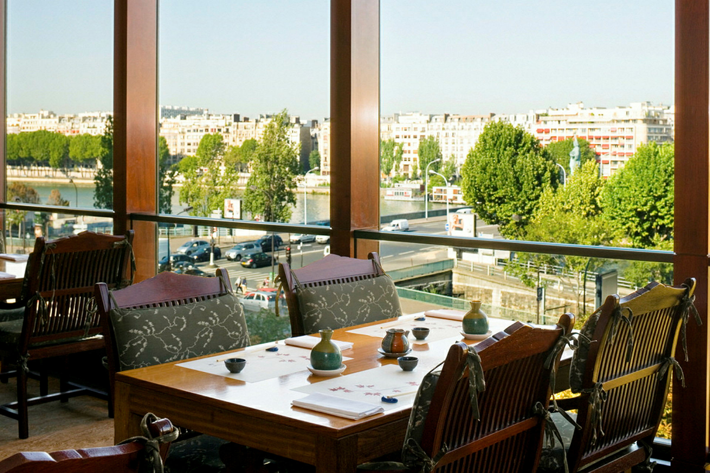 En restaurant med bord og stoler nær et stort vindu med utsikt over byen og elven. (AI-generert tekst)