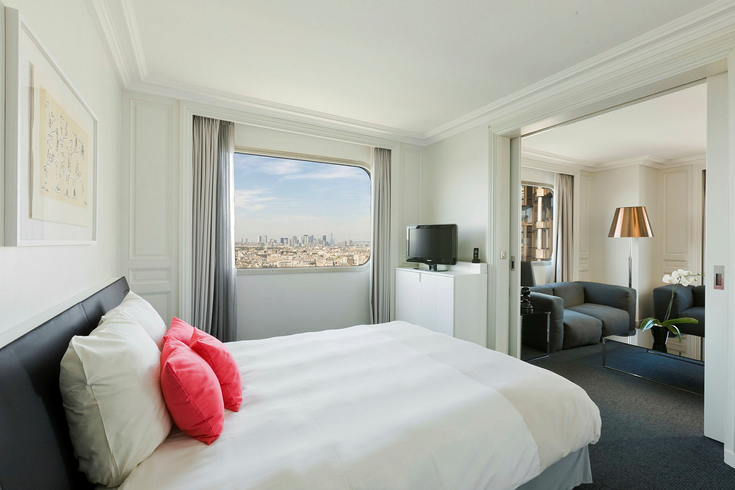 Et lyst og moderne hotellrom med en seng med hvite og rosa puter, vindu med utsikt over en by og en separat sittegruppe. (AI-generert tekst)