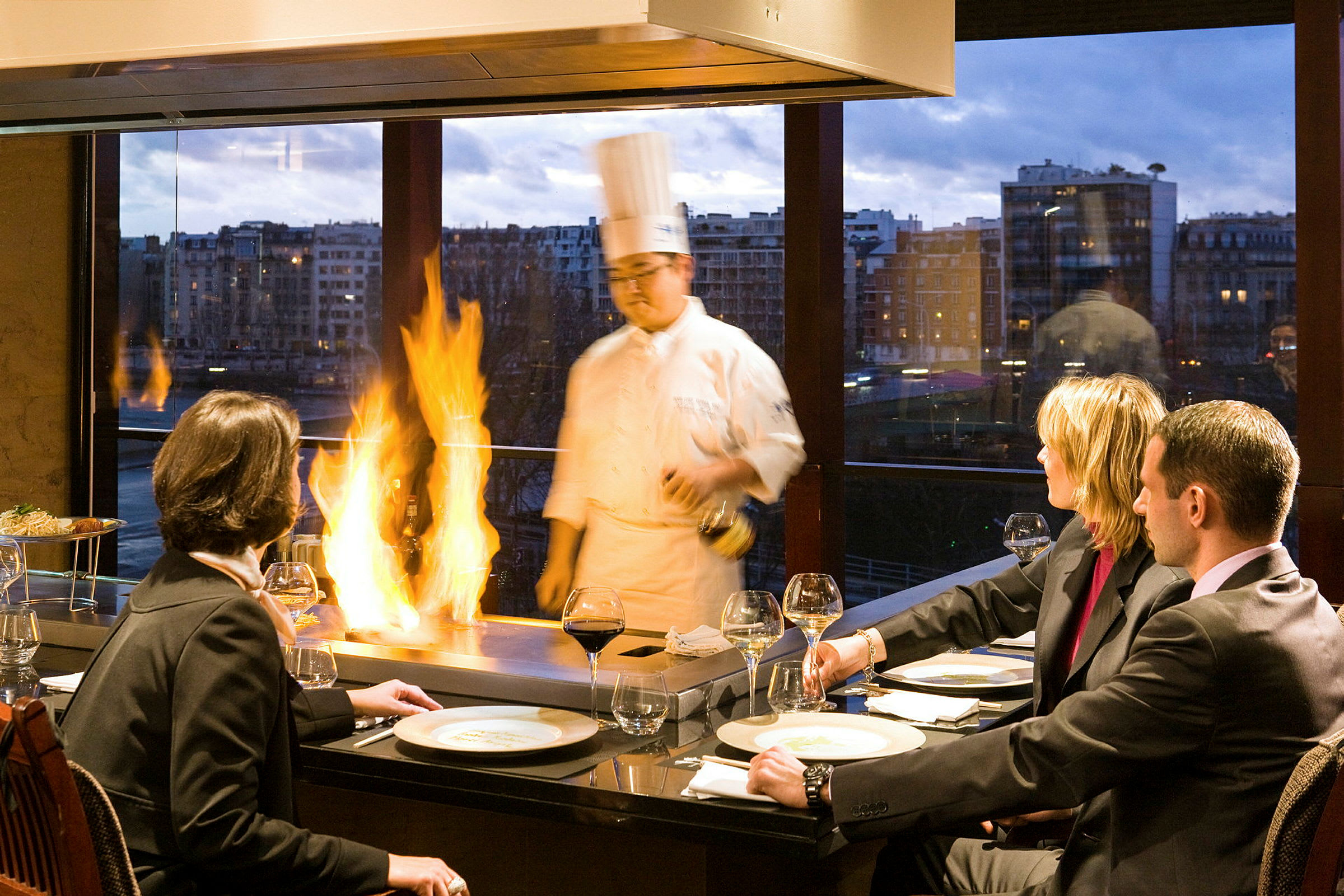 En gruppe mennesker sitter ved et bord og nyter et måltid mens kokken tilbereder mat med flammeeffekt i en restaurant med store vinduer og kveldutsikt. (AI-generert tekst)