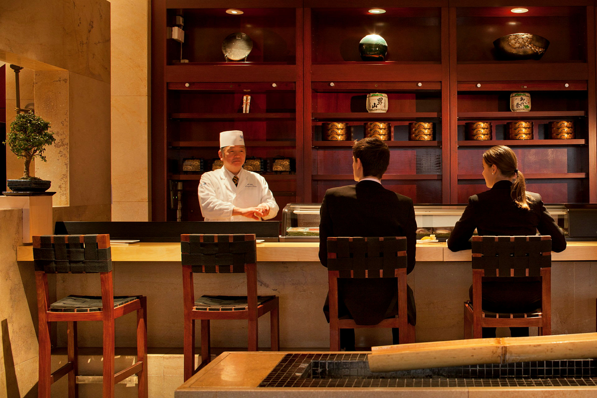 To personer sitter ved en sushi-bar med en kokk bak disken. (AI-generert tekst)