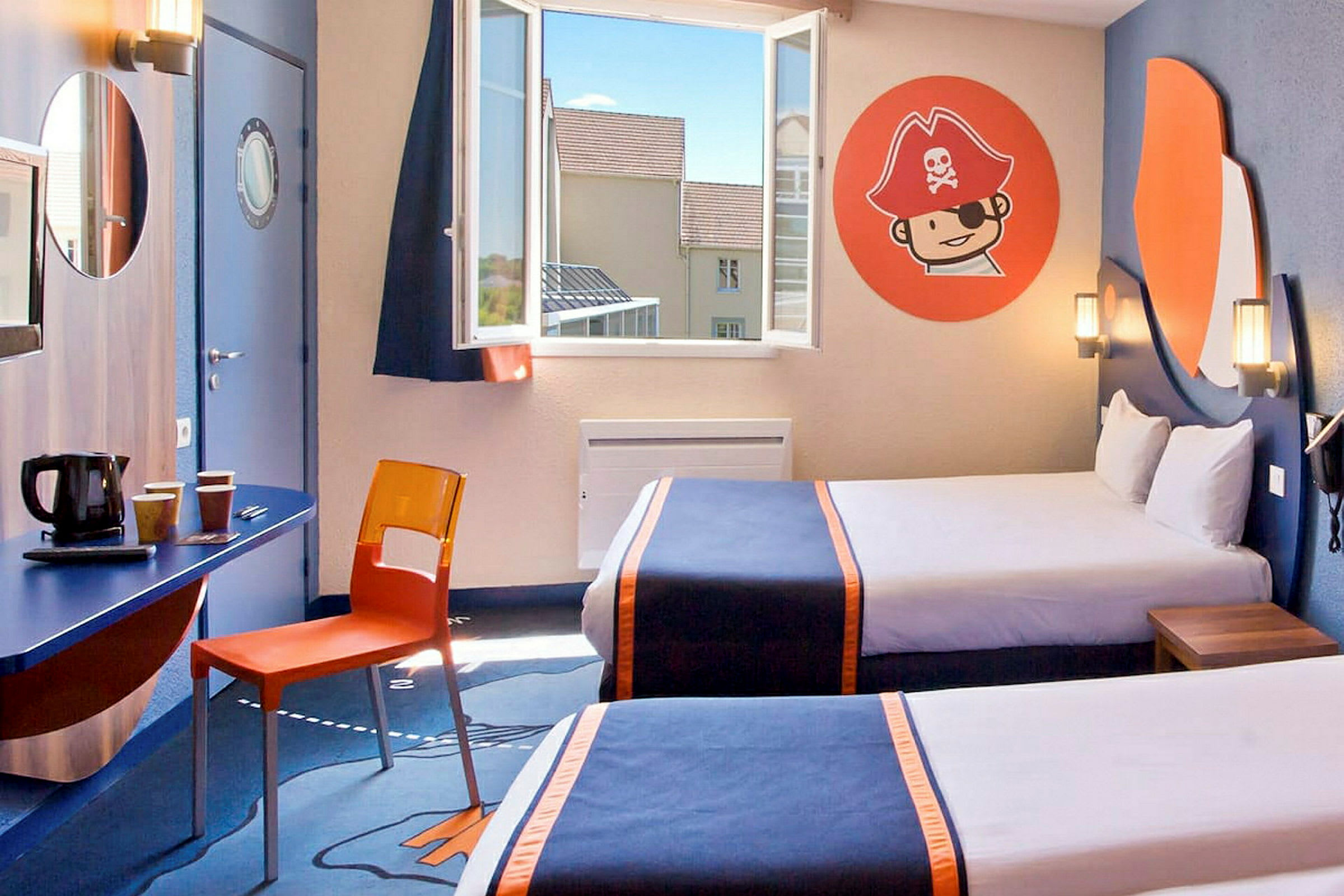 Et hotellrom med to senger dekket med mørkeblå sengetepper med oransje detaljer. Det er et vindu med åpen utsikt, en vegg med sjørøvertema på dekorative veggmalerier, samt et skrivebord med en oransje stol og kaffetrakter under et rundt speil. (AI-generert tekst)