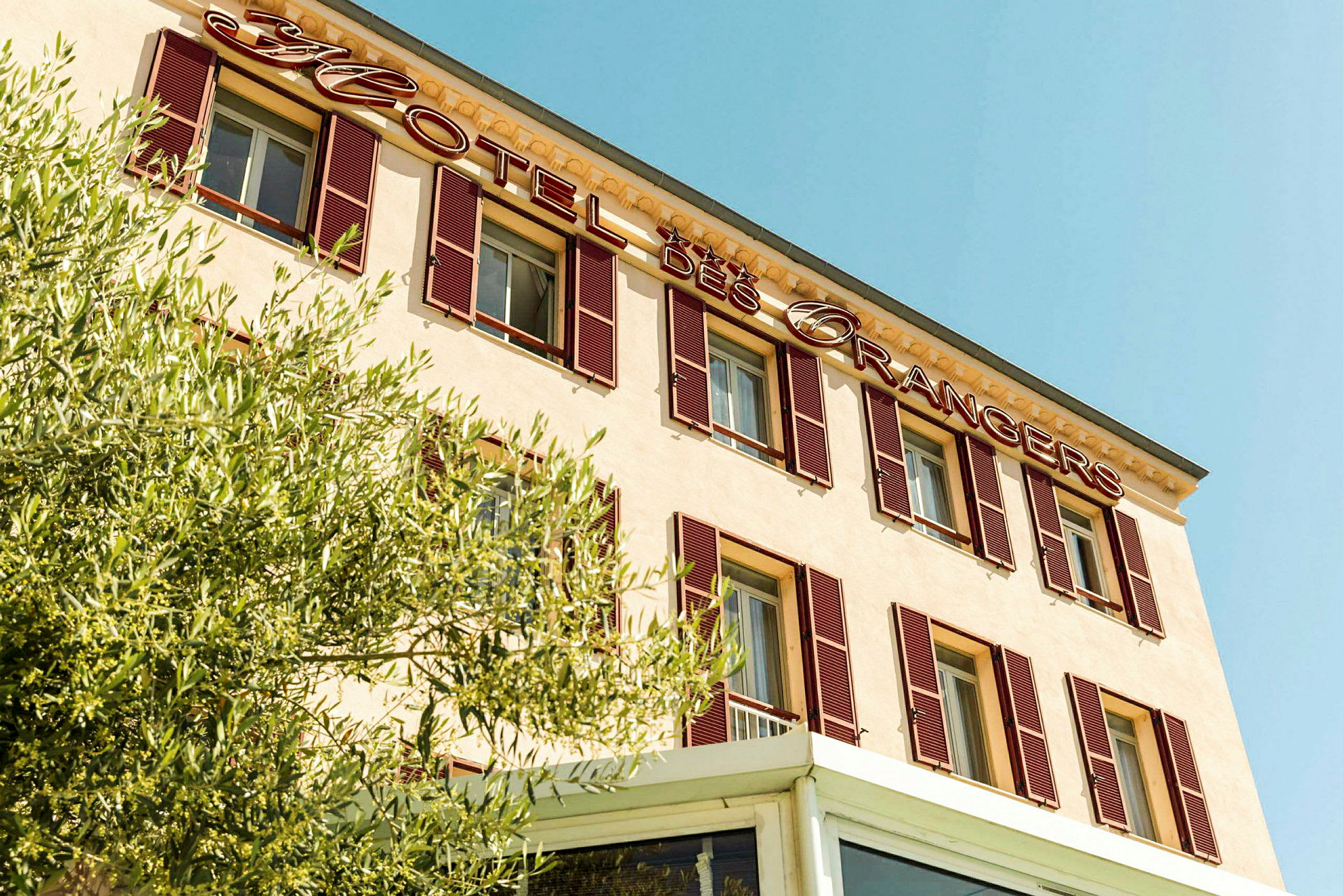 Fasade av hotellbygning med skiltet 'Hotel des Orangers' og grøntområder i forgrunnen. (AI-generert tekst)