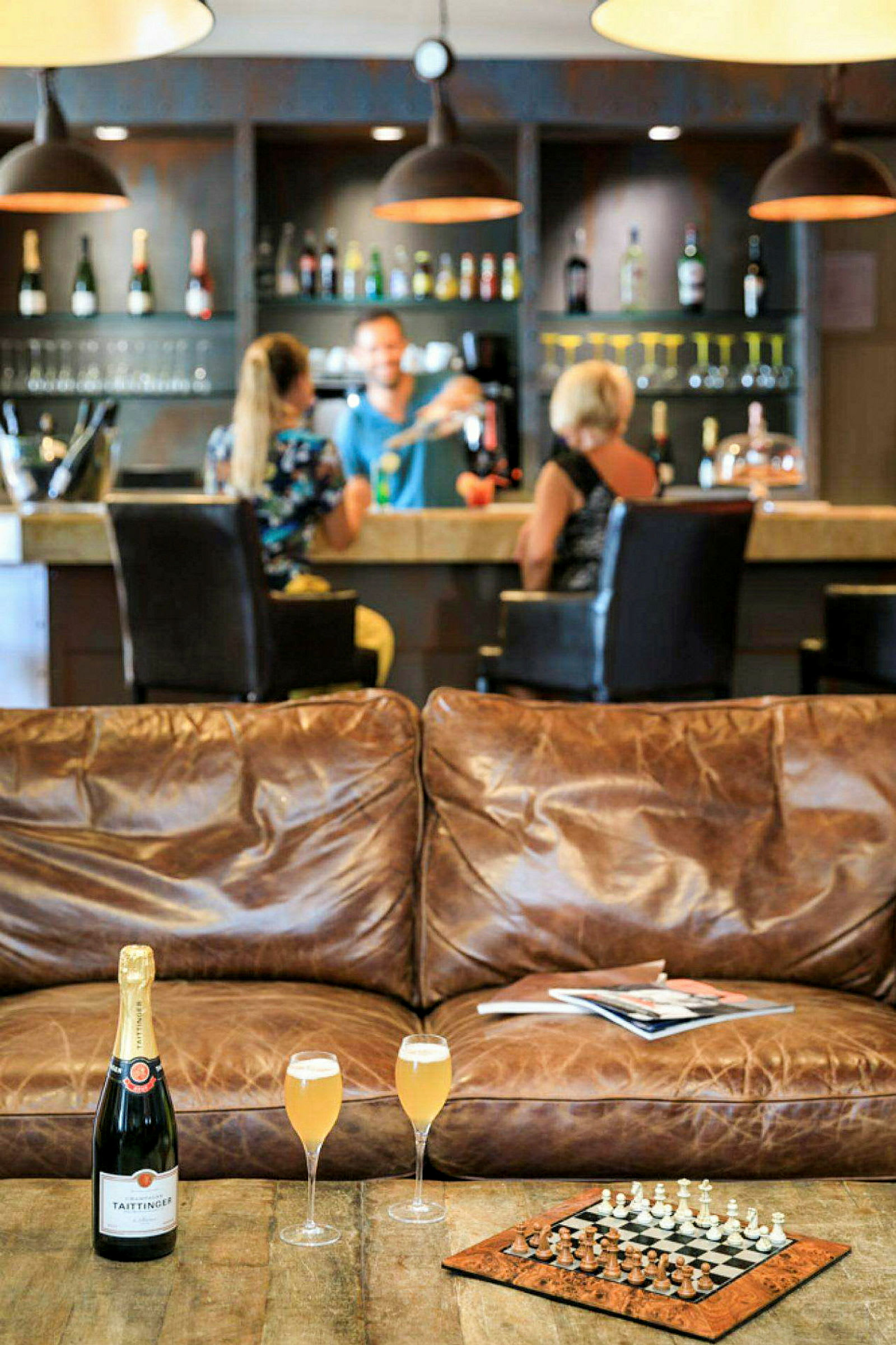 Interiørbilde av en bar med flasker i bakgrunnen, en skinnsofa, et sjakkspill og et bord med champagneglass i forgrunnen. (AI-generert tekst)
