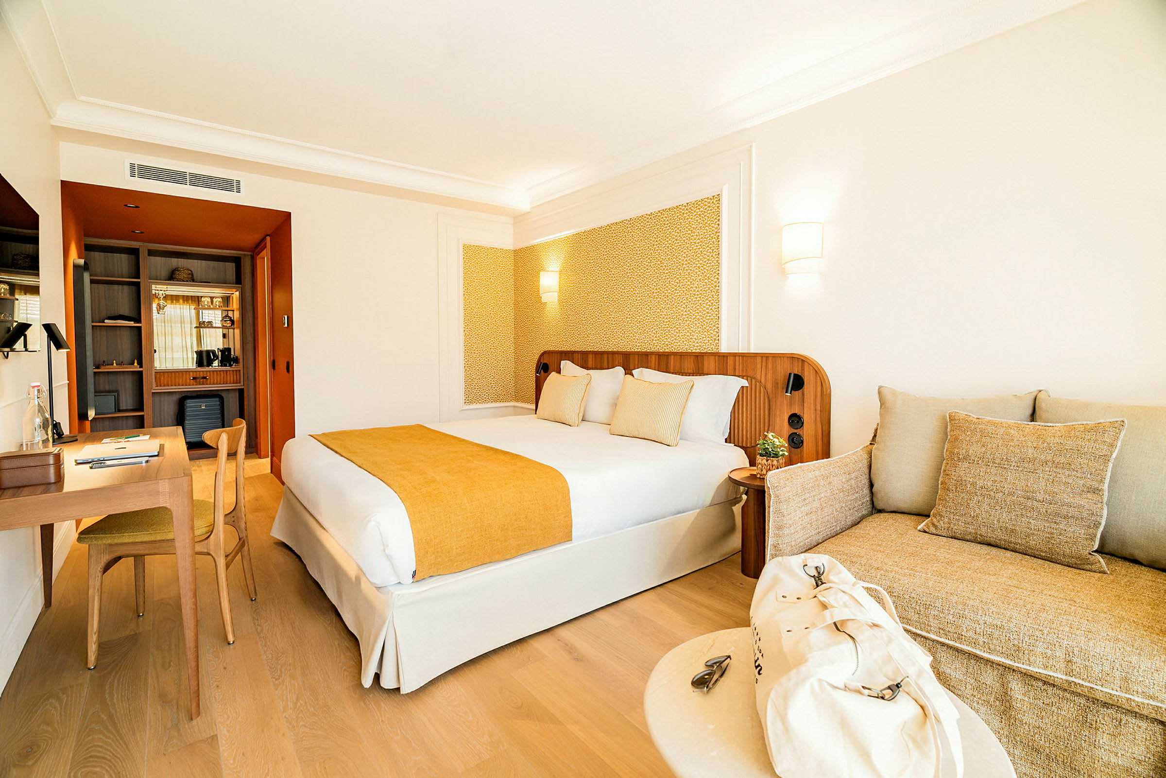 En hotellrominteriør med en dobbeltseng med gul sengeteppe og puter, et skrivebord med stol til venstre og en sofa med beige puter til høyre. (AI-generert tekst)