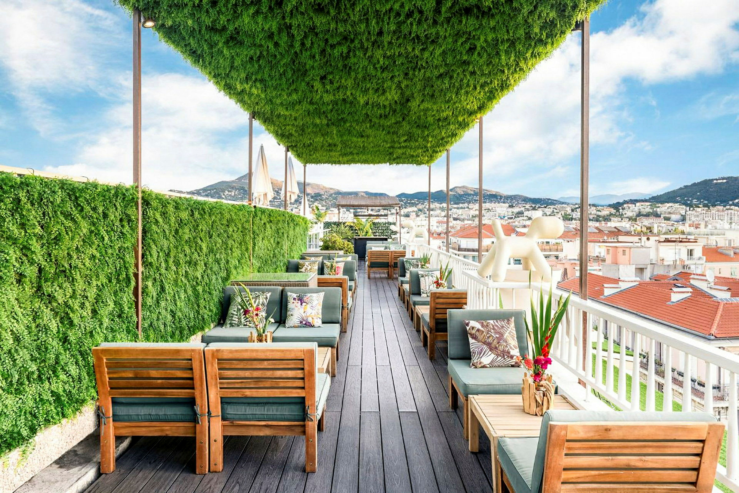 En takterrasse med grønt tak og planter, møblert med tremøbler og bord, med utsikt over hustak og fjell. (AI-generert tekst)
