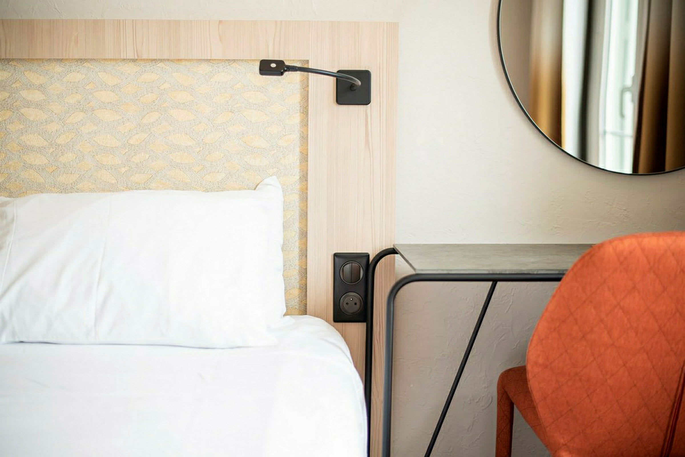 En hotellseng med en hvit pute og et nattbord ved siden av, samt en oransje stol foran et bord og et rundt speil på veggen. (AI-generert tekst)