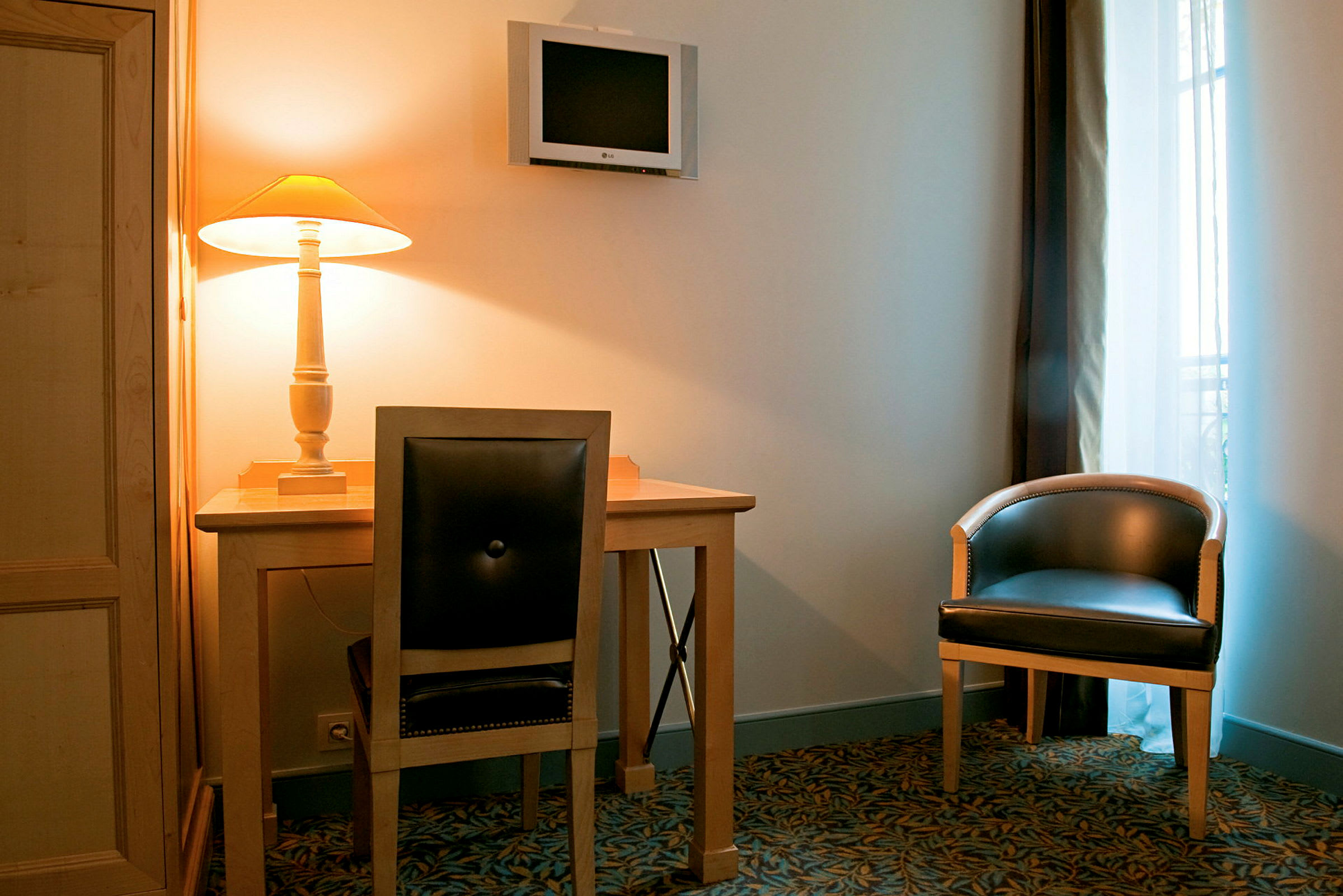 En innredning med et skrivebord og stol, en veggmontert TV og en lenestol ved siden av et vindu. Det står en tent bordlampe på skrivebordet. (AI-generert tekst)